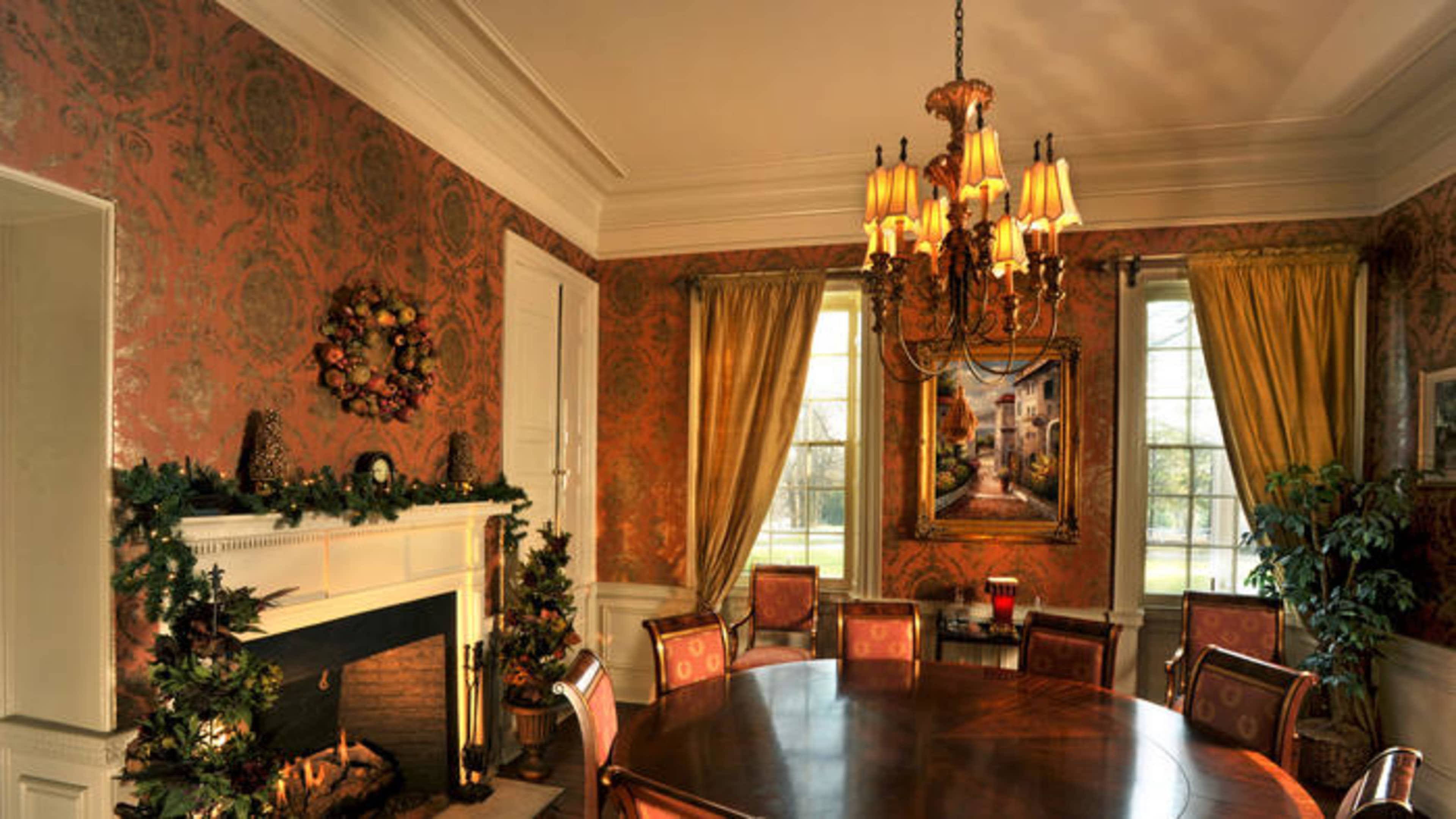 The dining room features a large round table surrounded by upholstered chairs, with a decorative fireplace and a painting on the wall amid festive holiday decorations.
