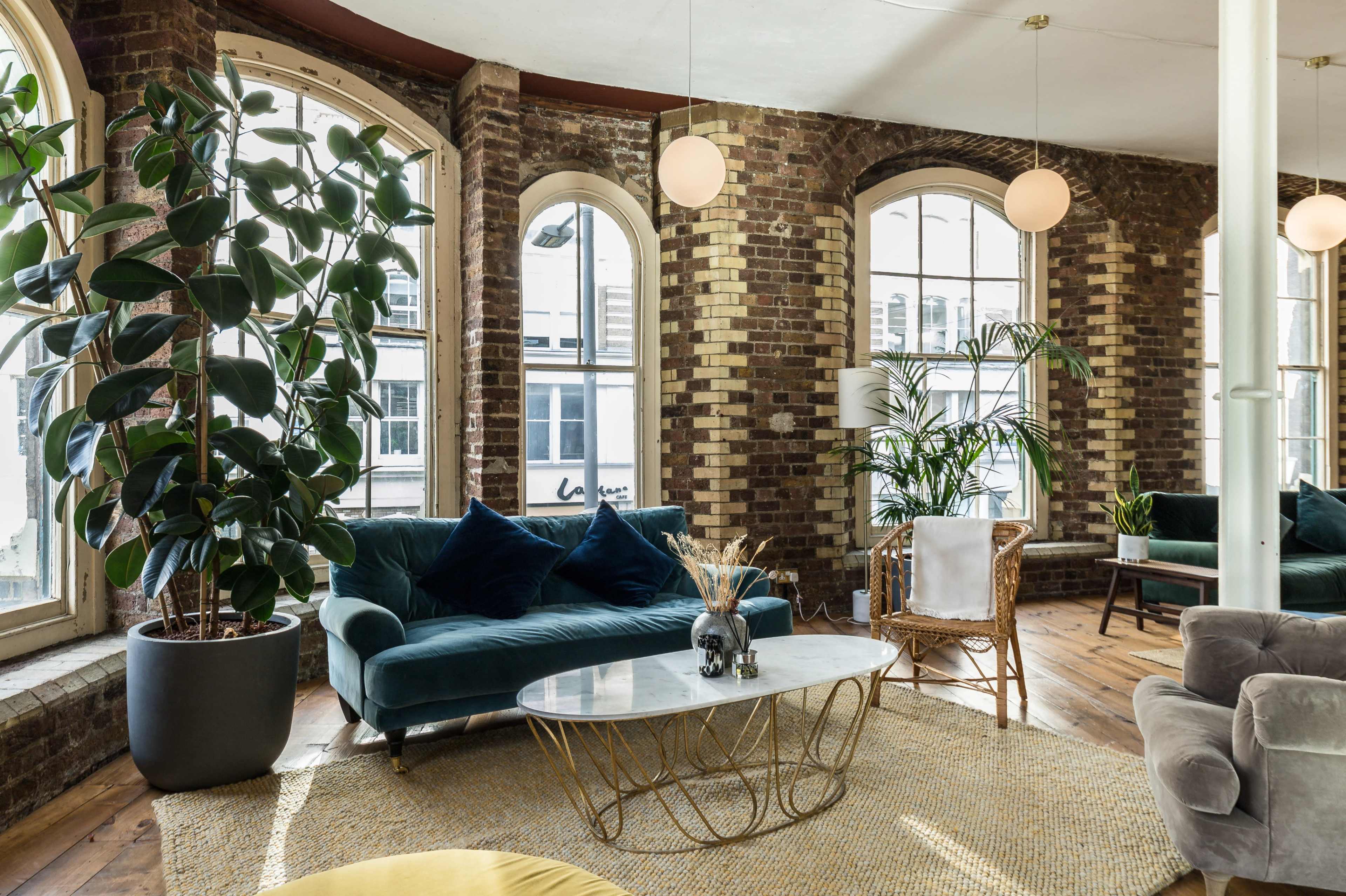 A modern living room features a velvet sofa, a glass-top coffee table, and large windows with brick walls, surrounded by potted plants.