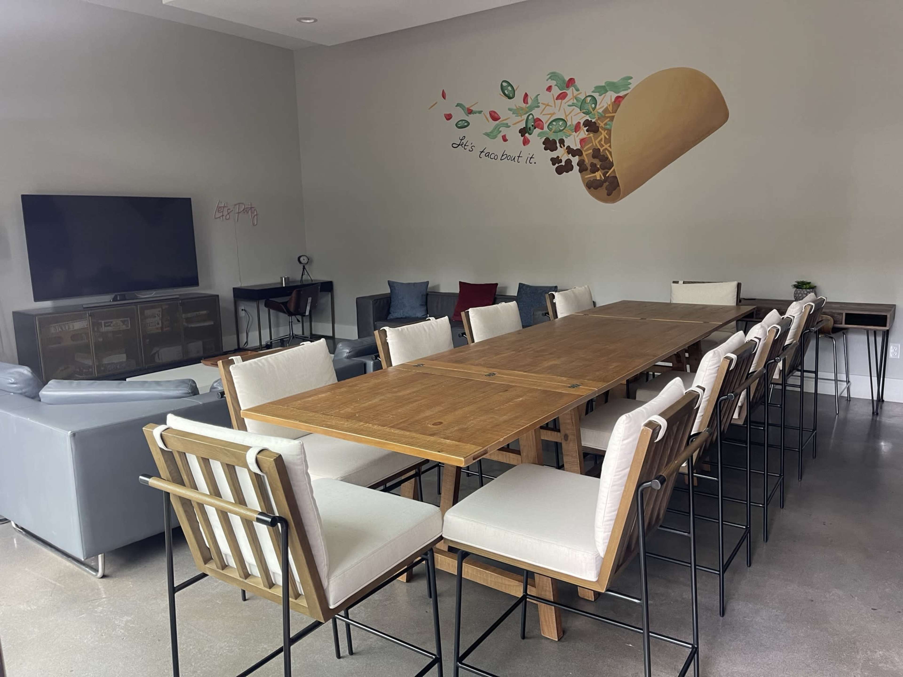 A spacious room with a long wooden dining table surrounded by high-backed chairs, a sectional couch in the corner, a television, and a mural of a taco on the wall.