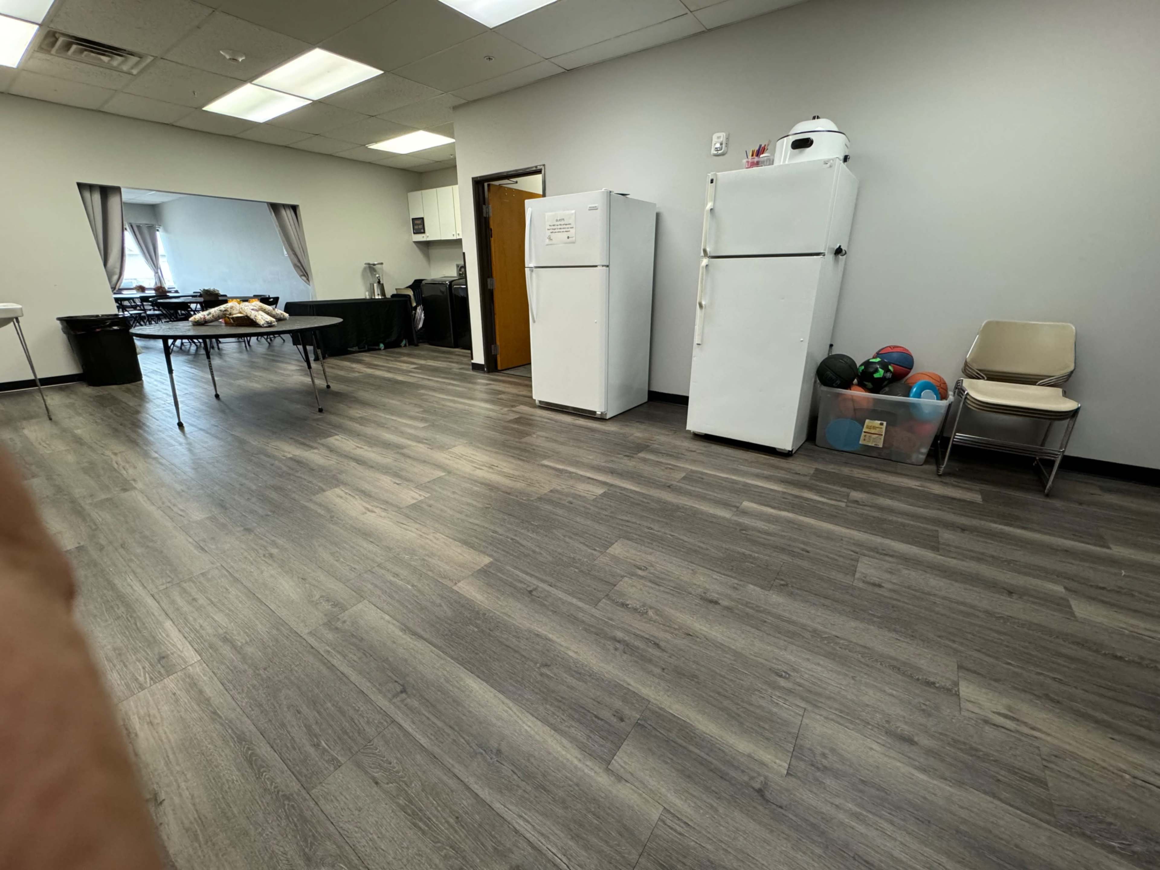 A room with two white refrigerators, a table covered with items, chairs, and exercise equipment in a corner.