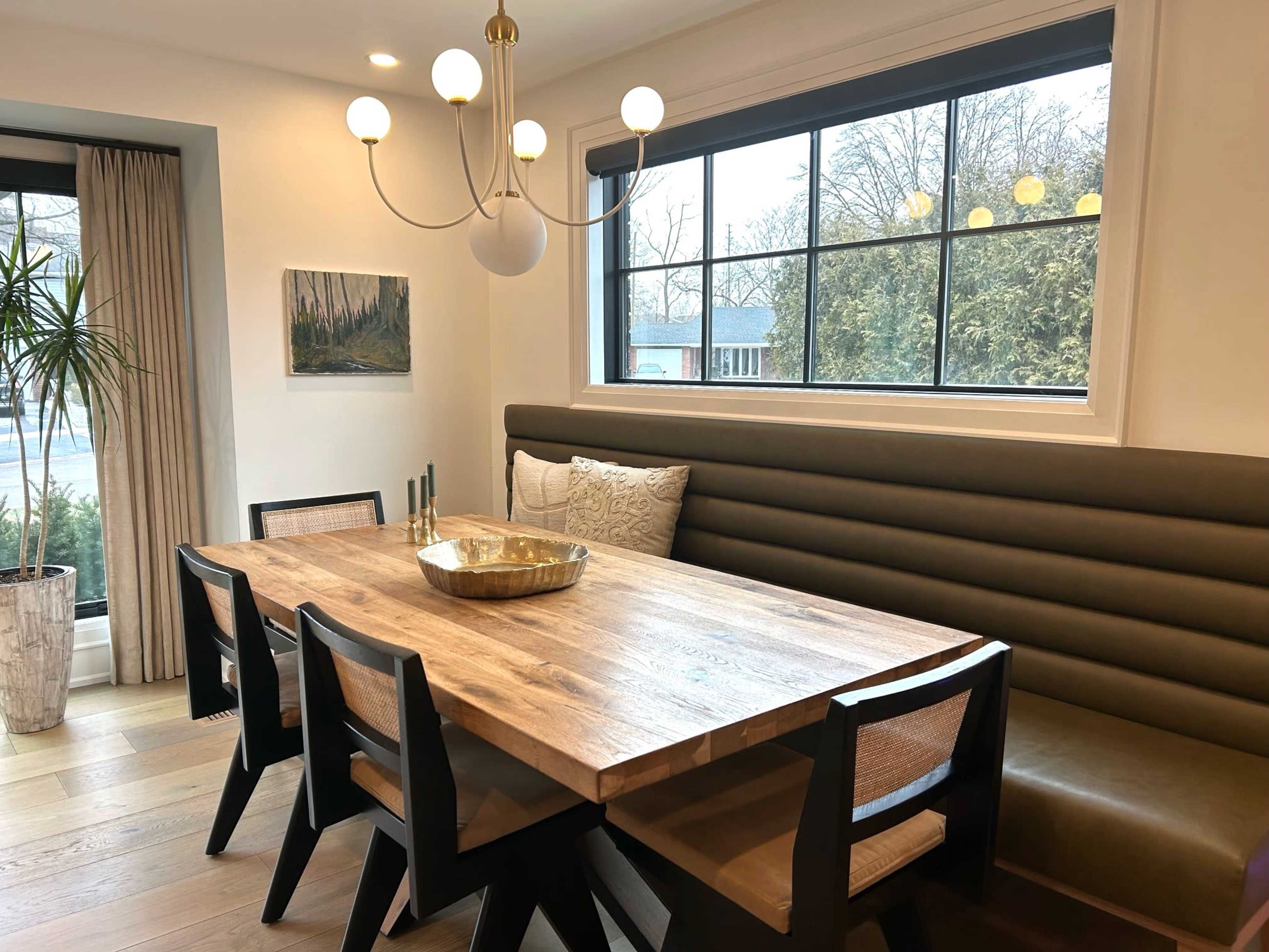 A modern dining area features a wooden table surrounded by black chairs, a green banquette, and a chandelier above, with large windows providing natural light.