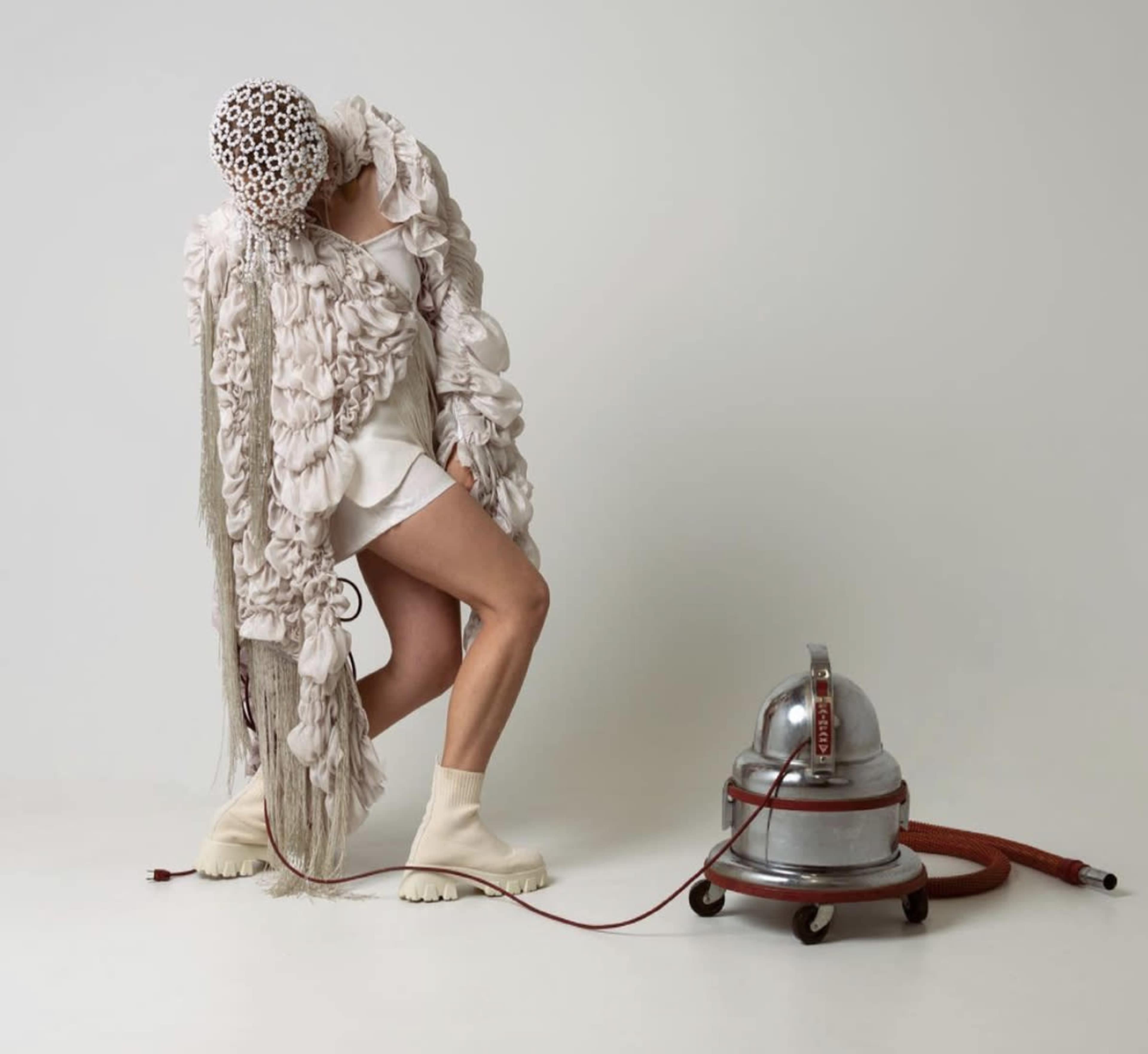 A person in an elaborate textured outfit interacts with a vintage vacuum cleaner in a minimalist setting.