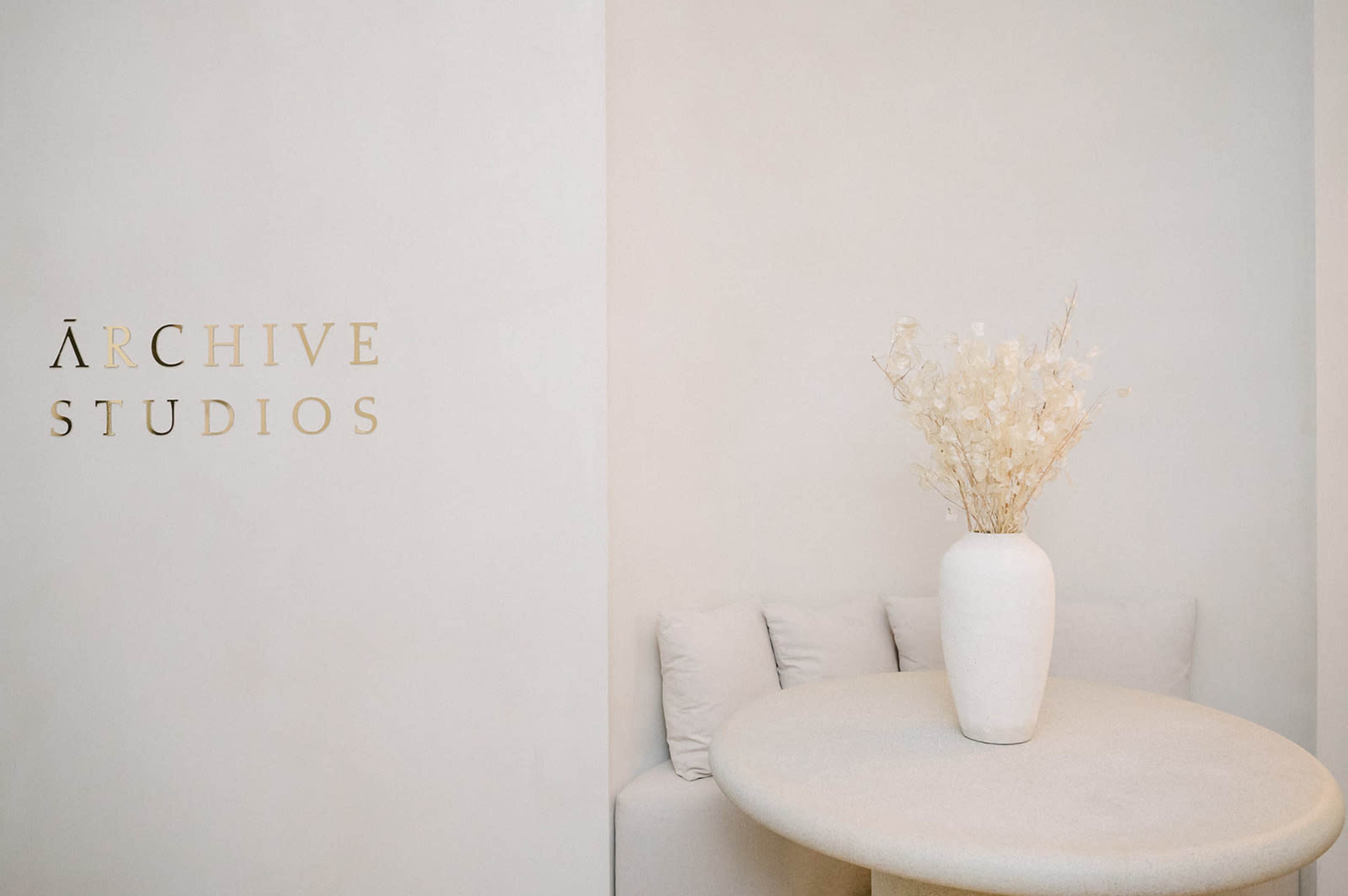 A minimalist interior features a round table with a vase of dried flowers against a light-colored wall displaying the words "Archive Studios."