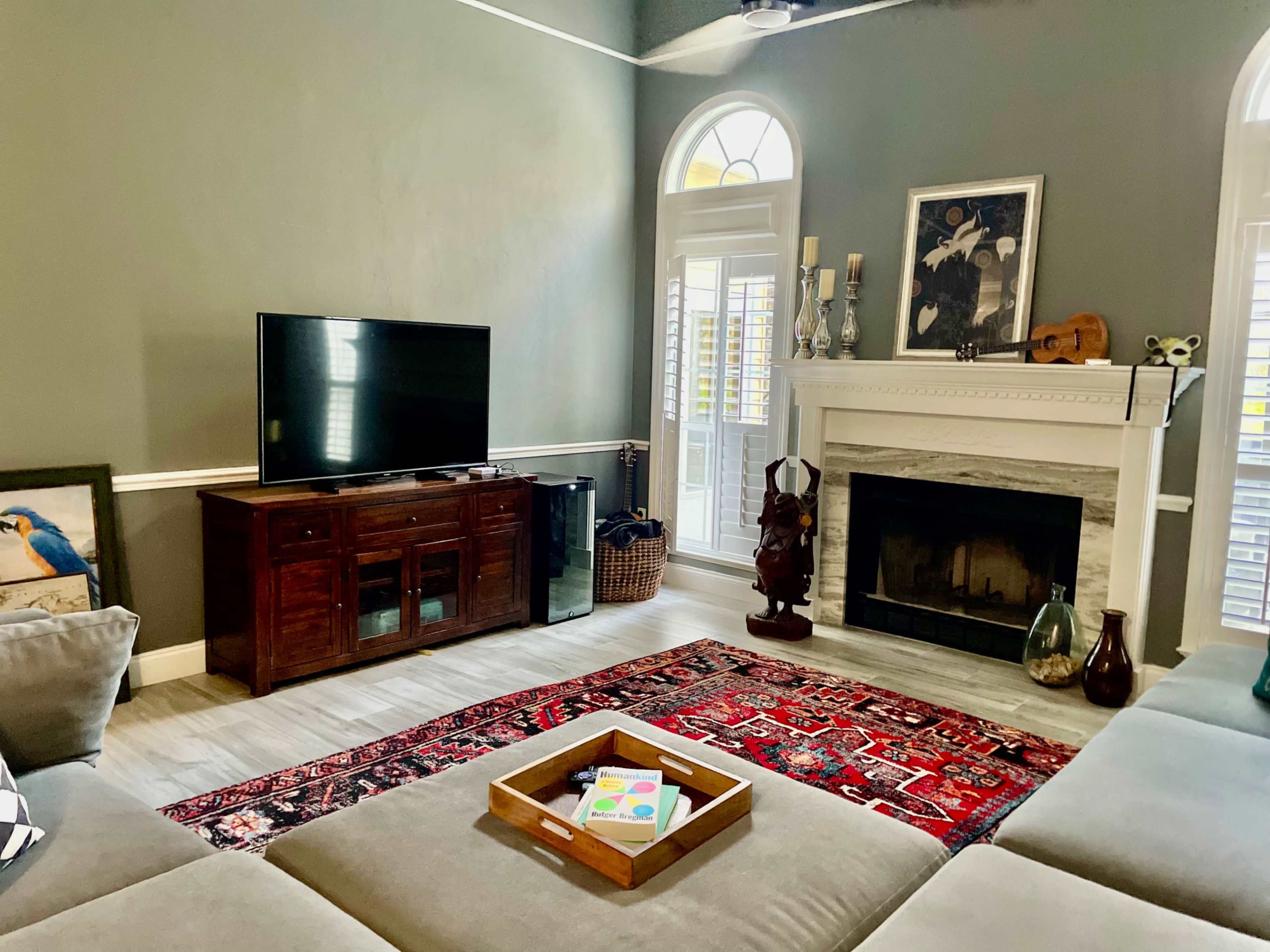 A living room features a television on a wooden cabinet, a stone fireplace, and a rug with a decorative pattern, all surrounded by large windows and natural light.