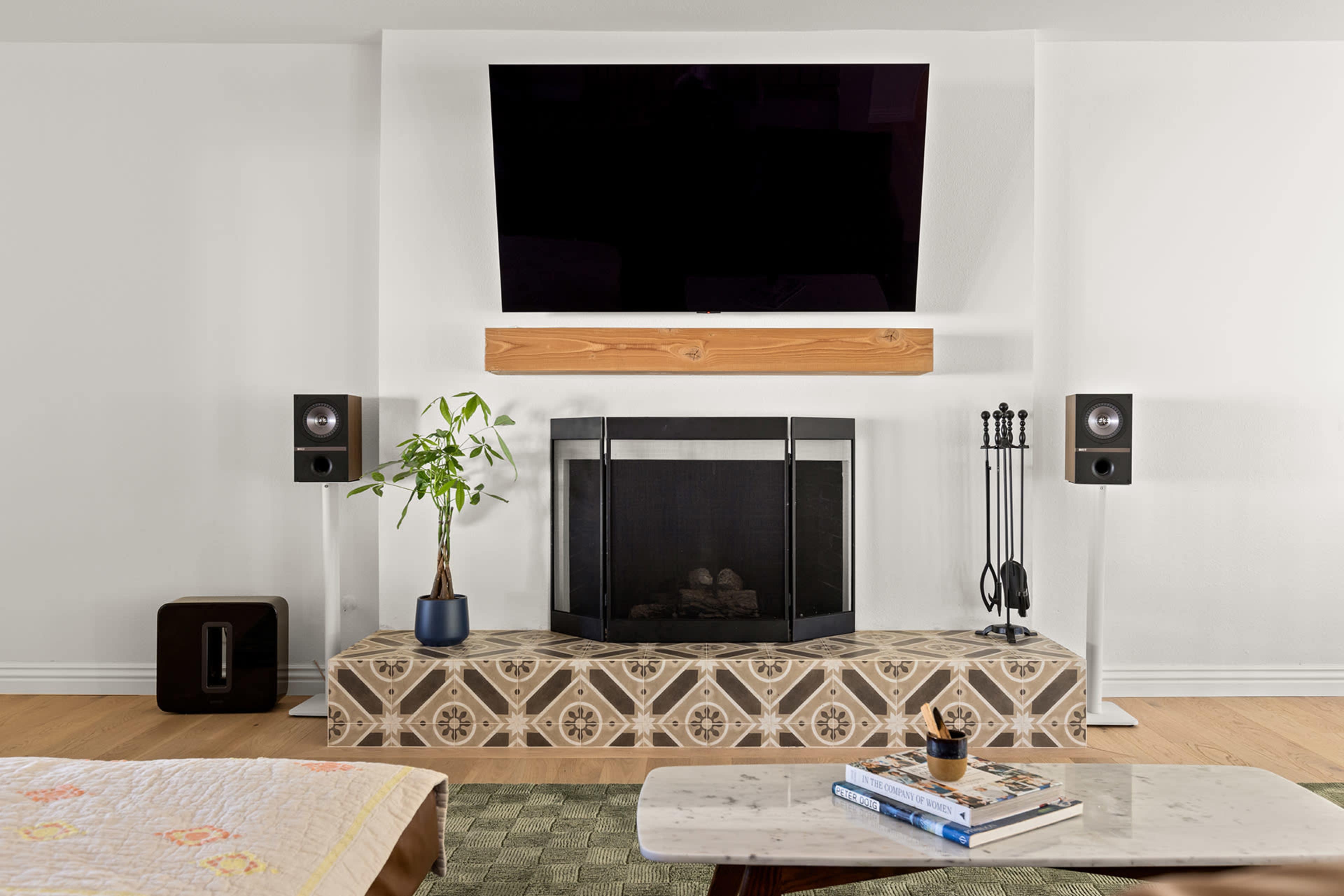 A modern living room with a wall-mounted television above a decorated fireplace, flanked by two speakers and a potted plant in front.
