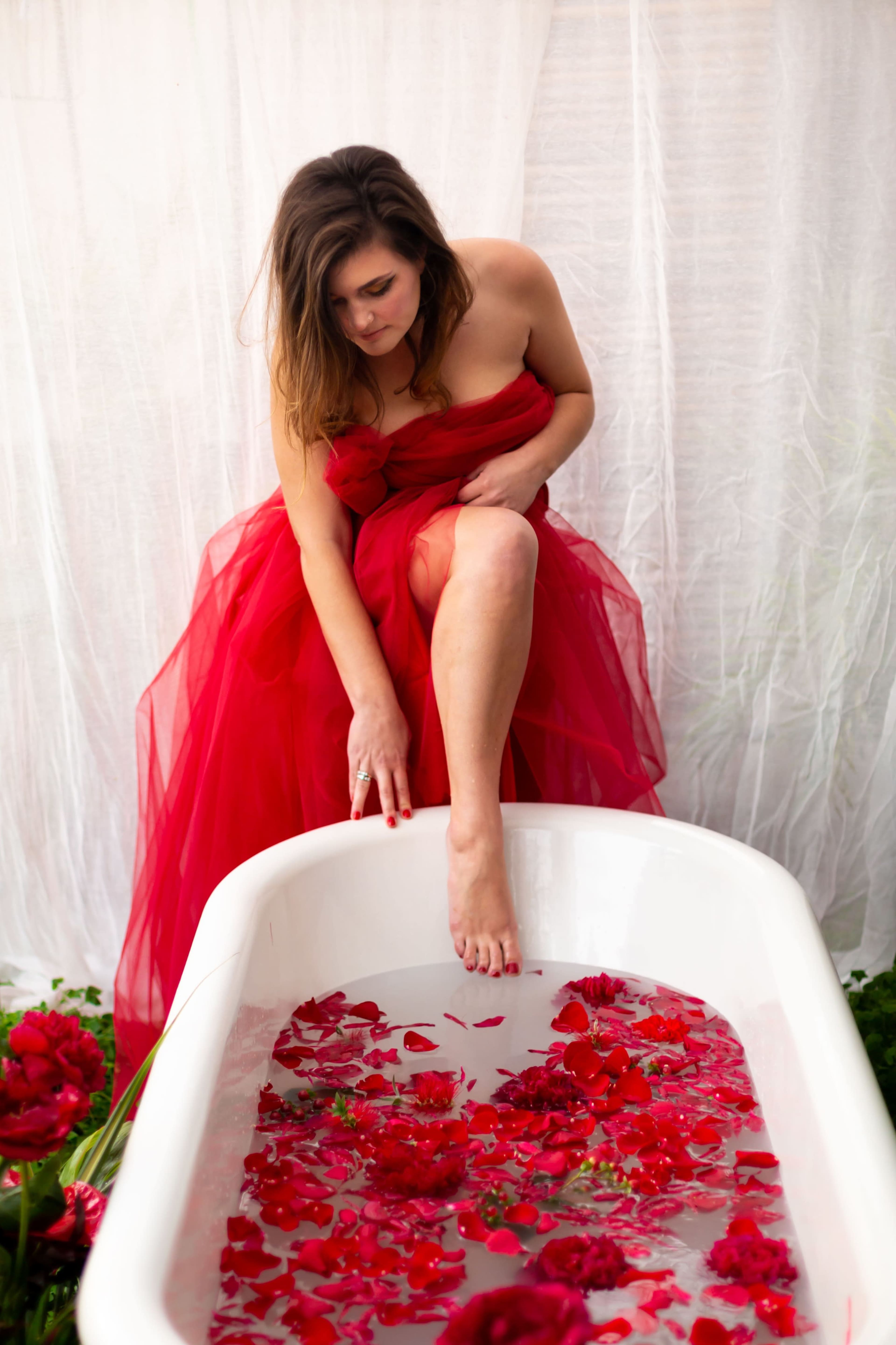 A woman in a red dress sits on the edge of a white bathtub filled with water and rose petals.
