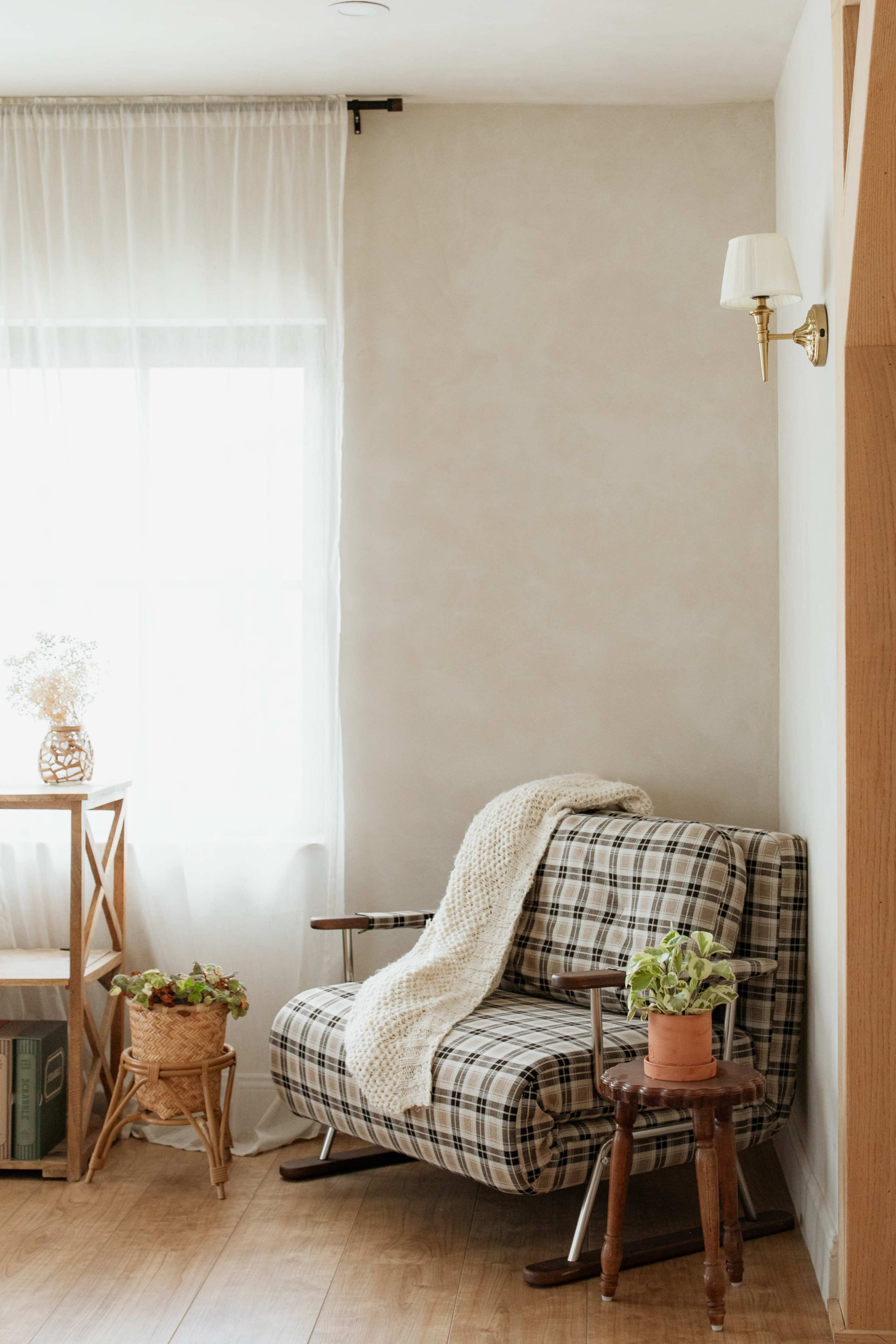 A checkered armchair is positioned next to a window, accompanied by a small table with plants and a wall sconce.