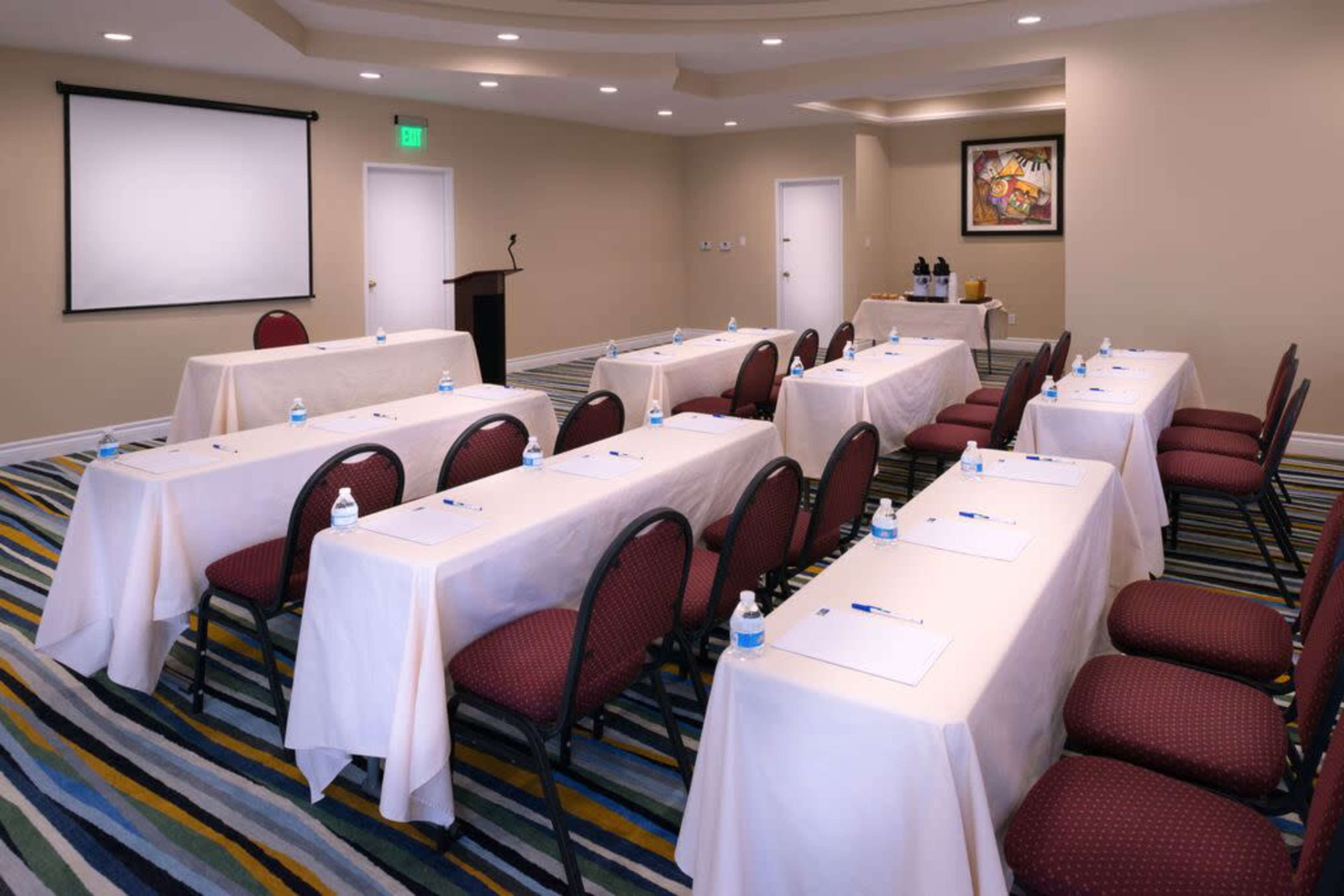 A meeting room arranged with tables covered in white cloth, each equipped with a notebook, pen, and water bottle, facing a projector screen.