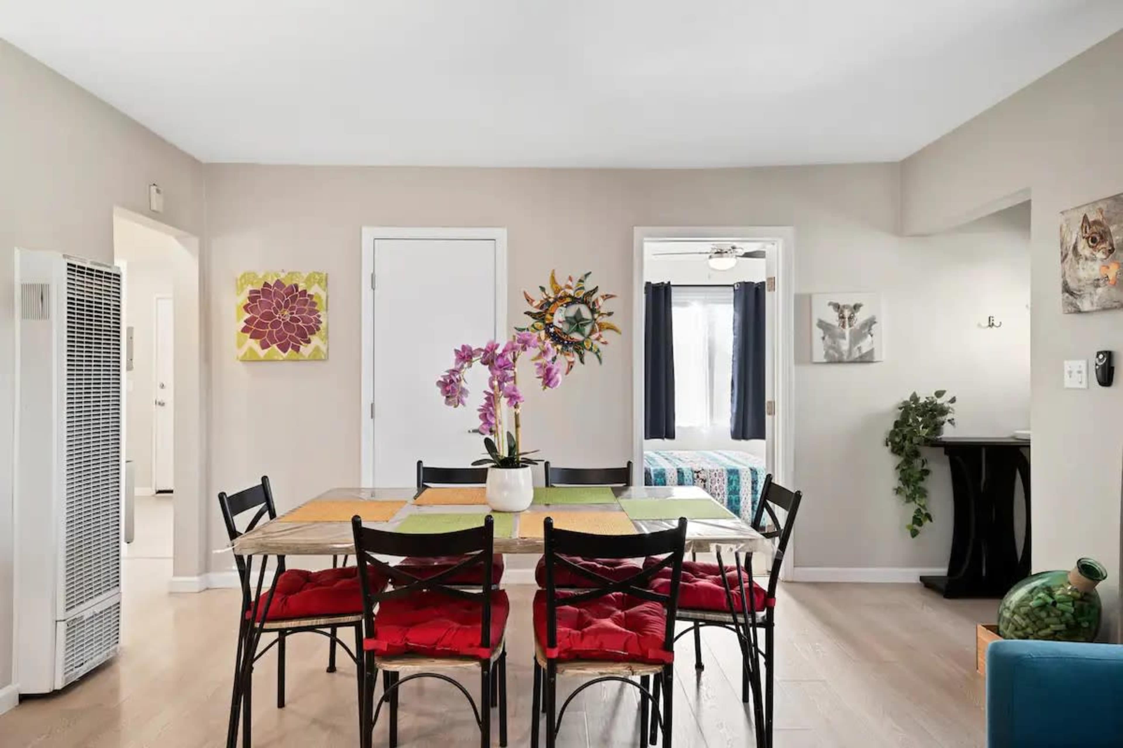 A dining area features a rectangular table with colorful placemats and black chairs, surrounded by light-colored walls and decorative wall art.