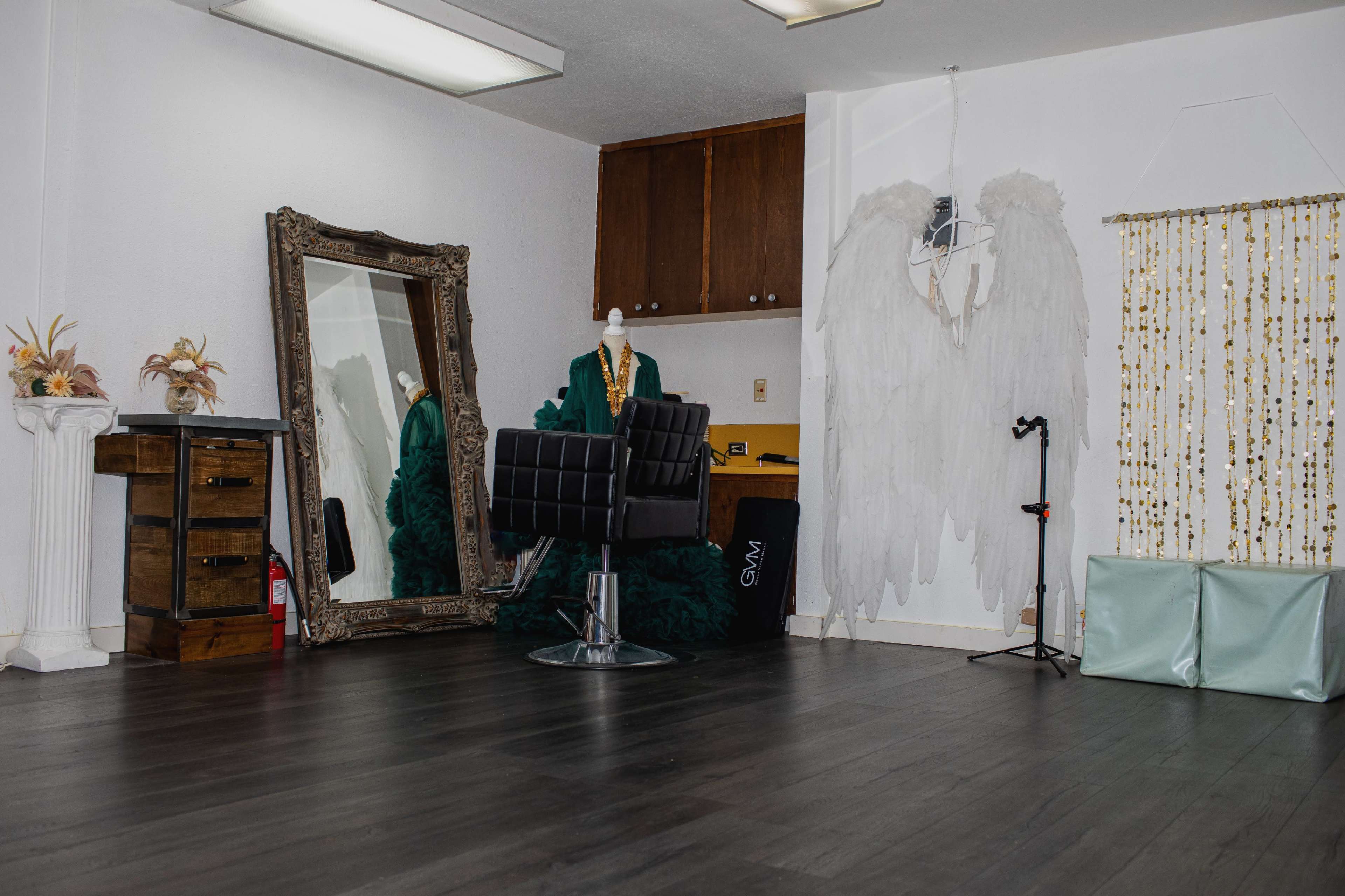 The image shows a spacious, minimalist room with a large mirror, a black salon chair, a mannequin wearing clothing, and decorative wings on the wall.