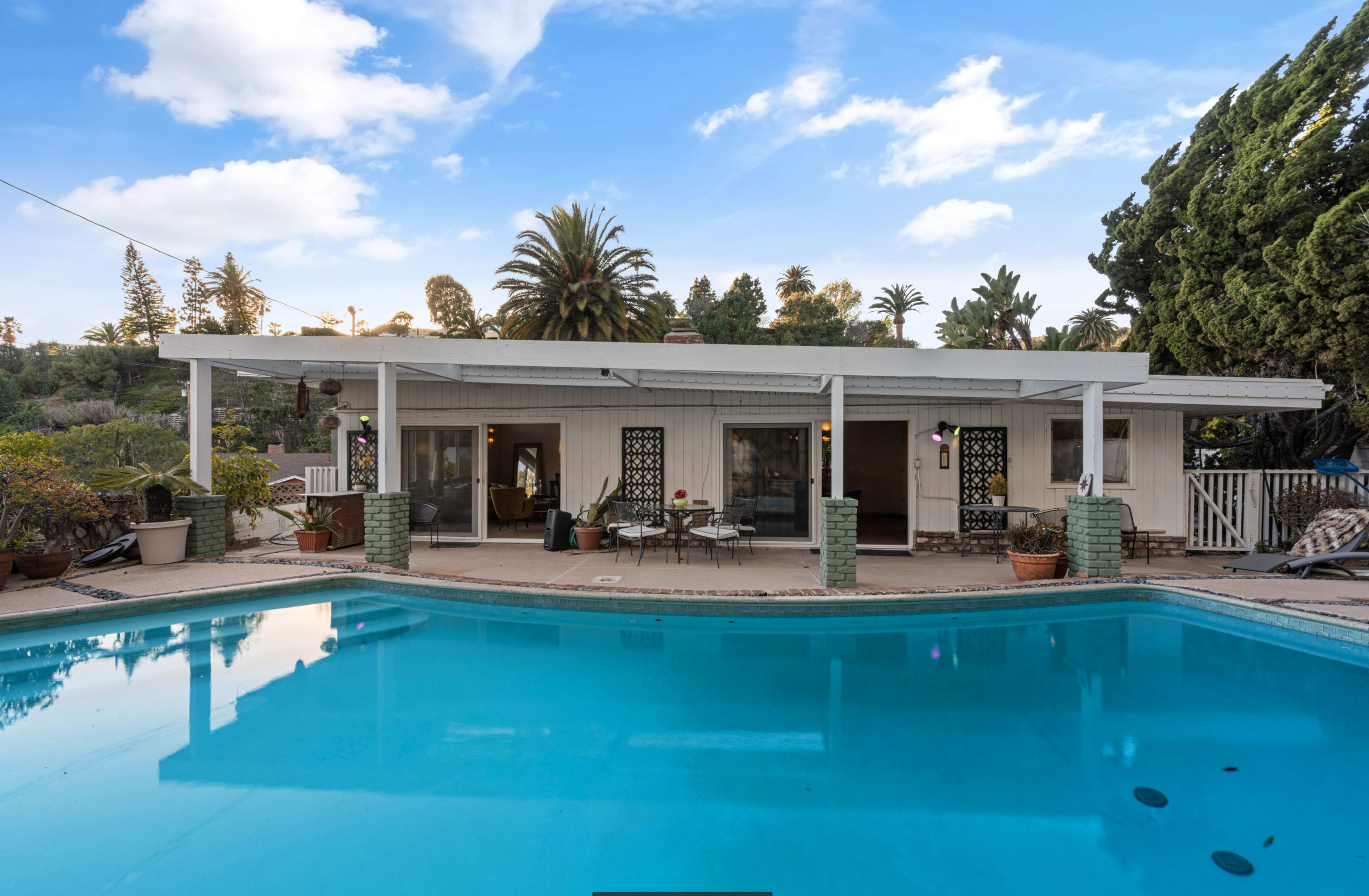 A single-story house with a pool in the foreground is surrounded by palm trees and greenery.
