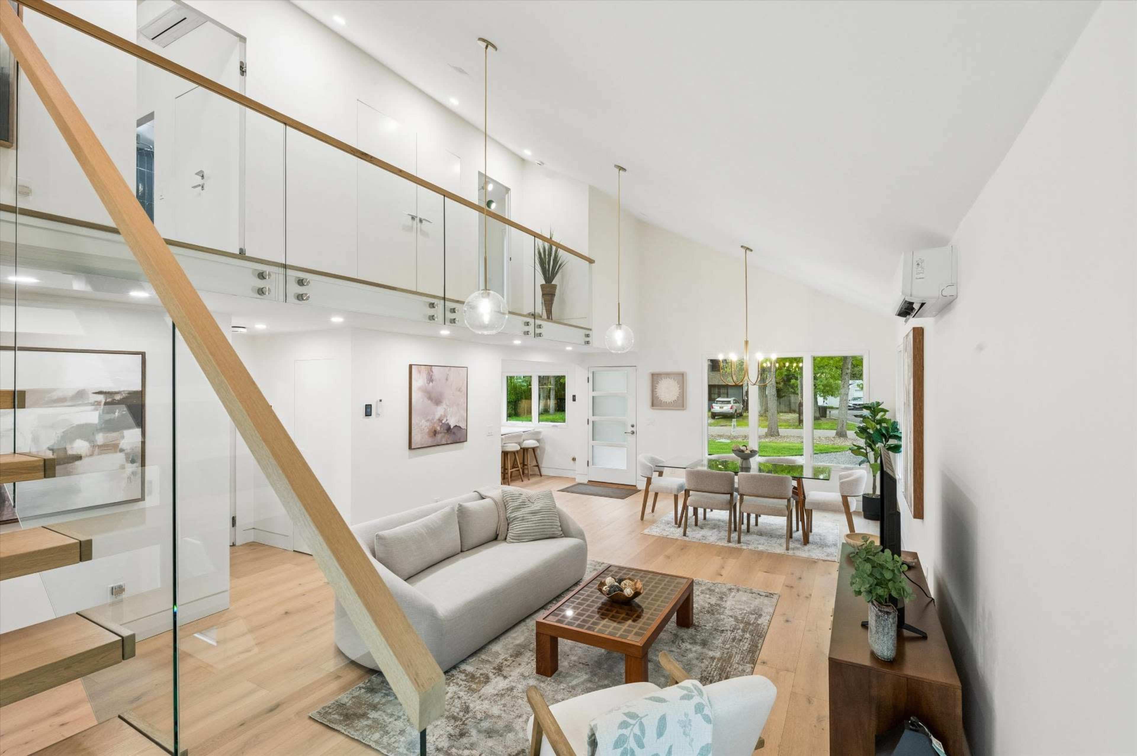 A modern living room features a light-colored sofa, wooden coffee table, and dining area with large windows allowing natural light to enter.