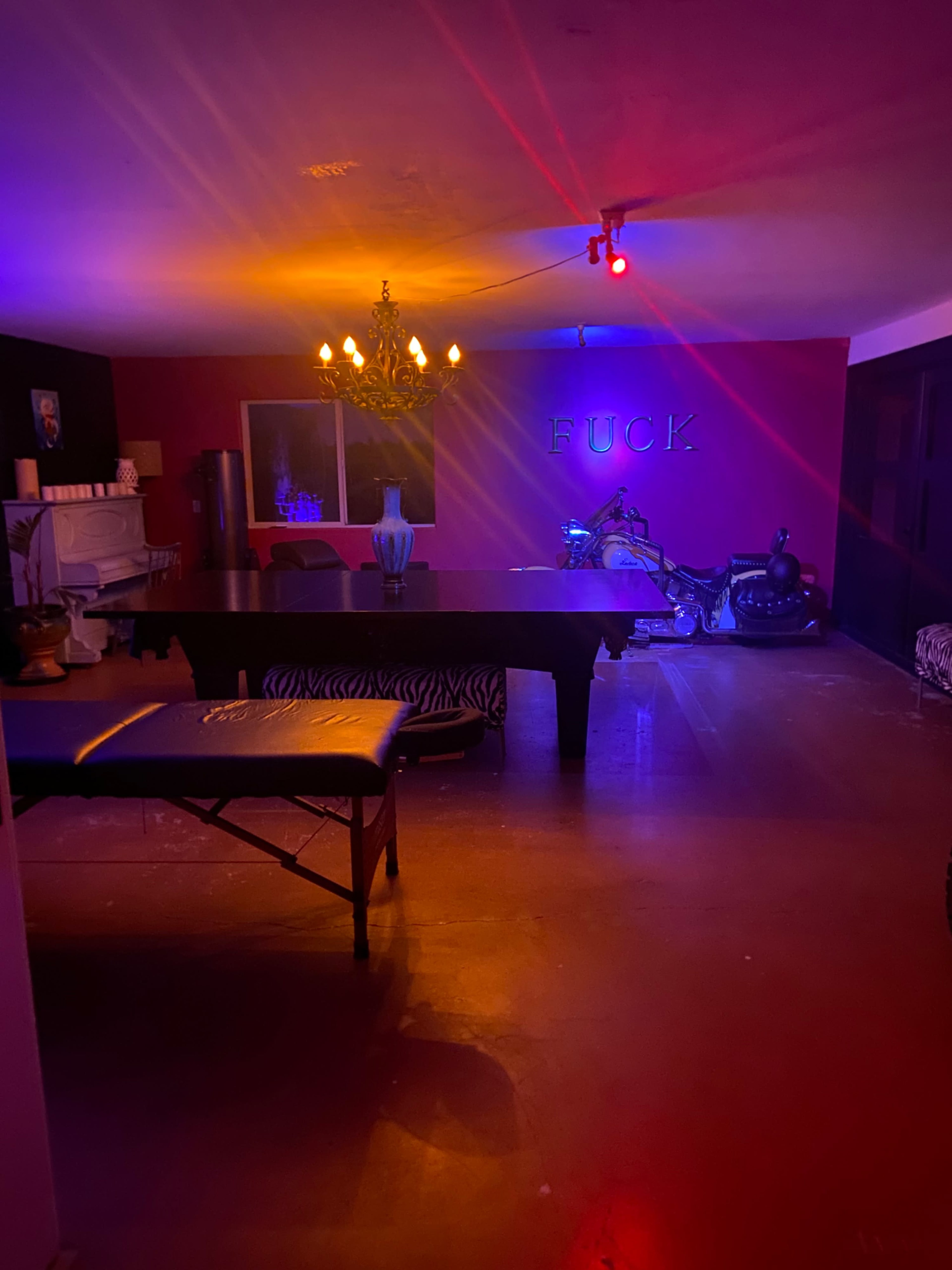 The room features a large table, a chandelier, and colorful lighting that illuminates the walls adorned with the word "FUCK."