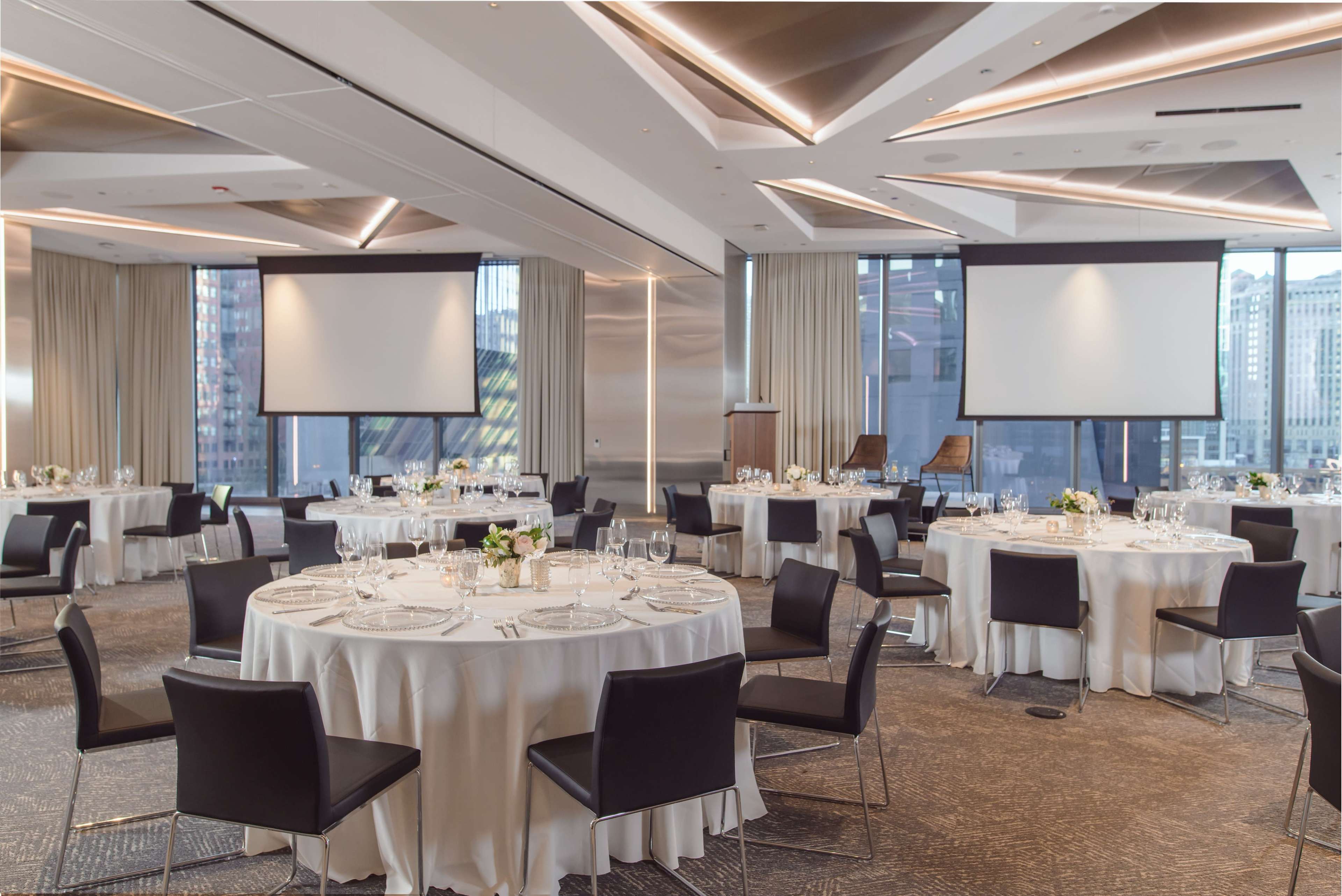 A modern event space features several round dining tables set with white tablecloths and black chairs, surrounded by large windows displaying an urban landscape.