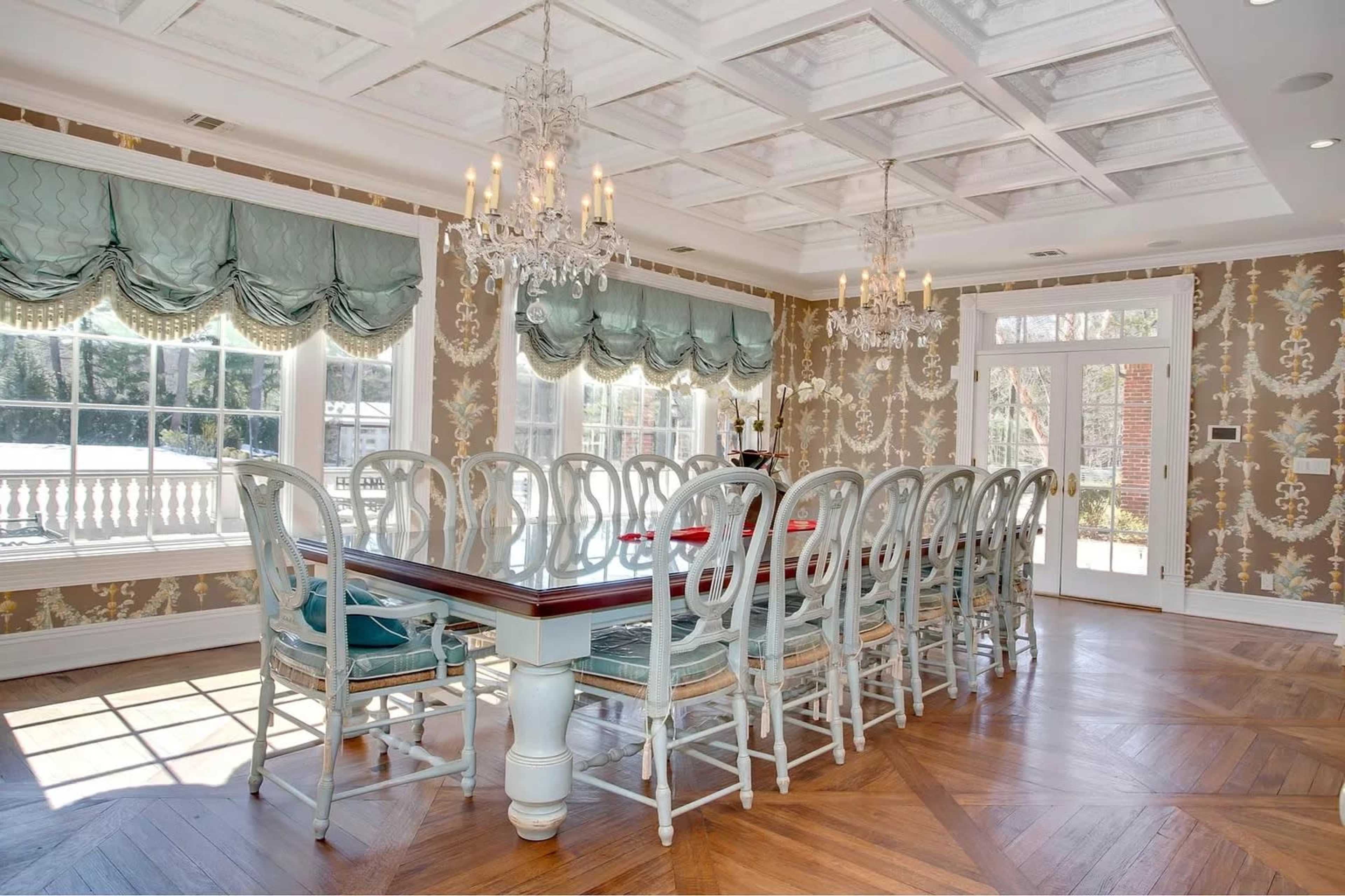 A spacious dining room with a large table surrounded by multiple chairs, featuring a coffered ceiling and elegant chandeliers.