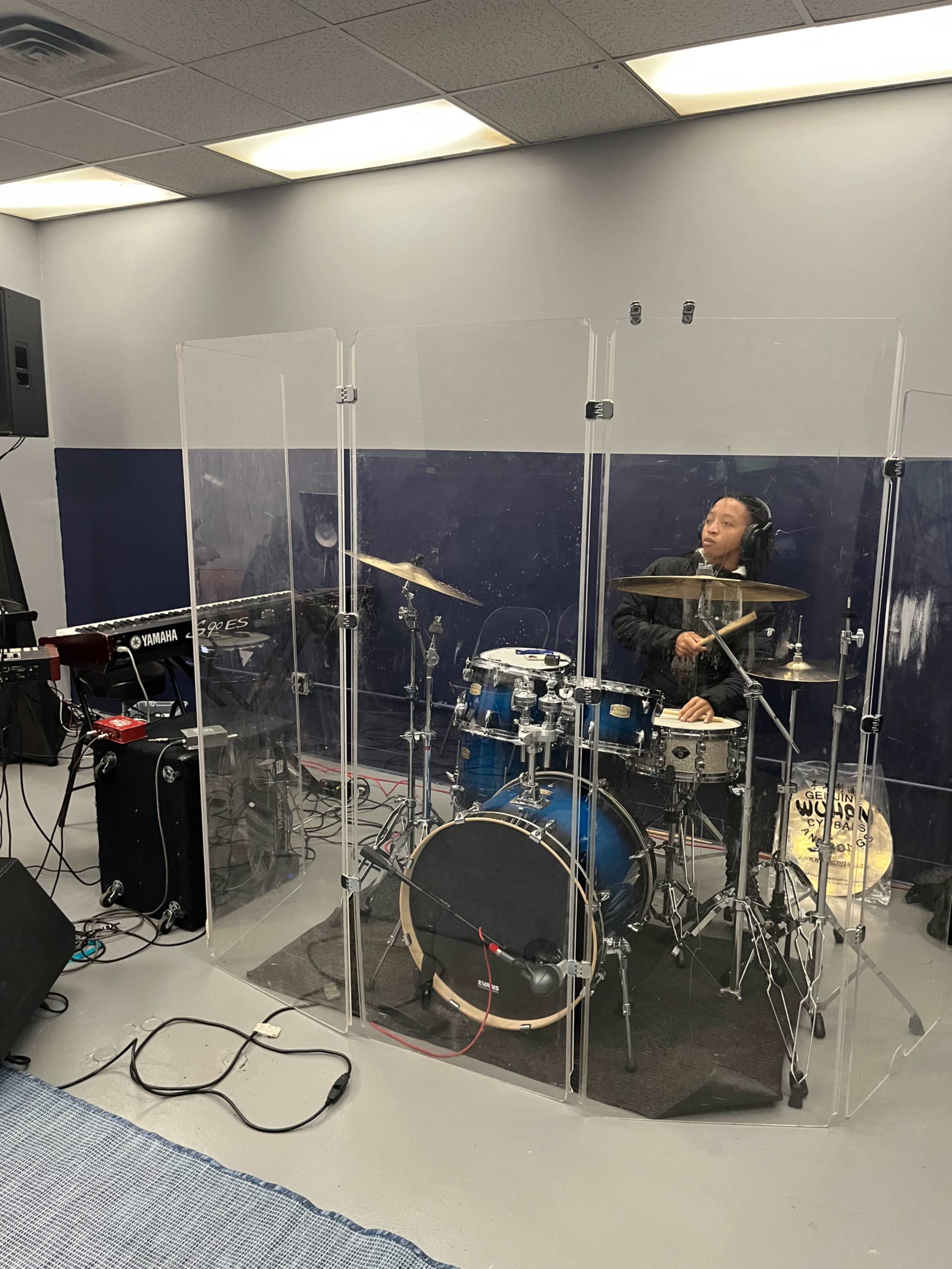 A drummer plays inside a transparent sound barrier while a keyboard is set up nearby in a rehearsal space.