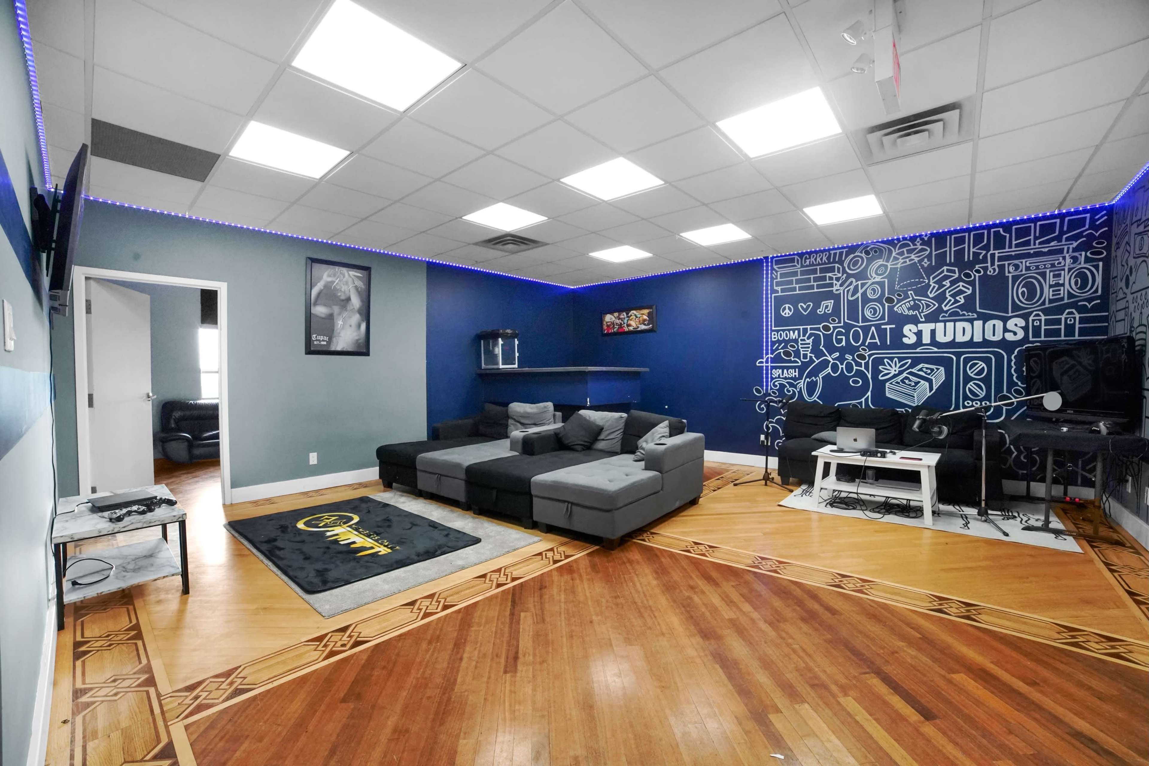A modern studio room with a sectional couch, a TV on the wall, and a workspace with a desk and computer equipment against a decorated blue wall.