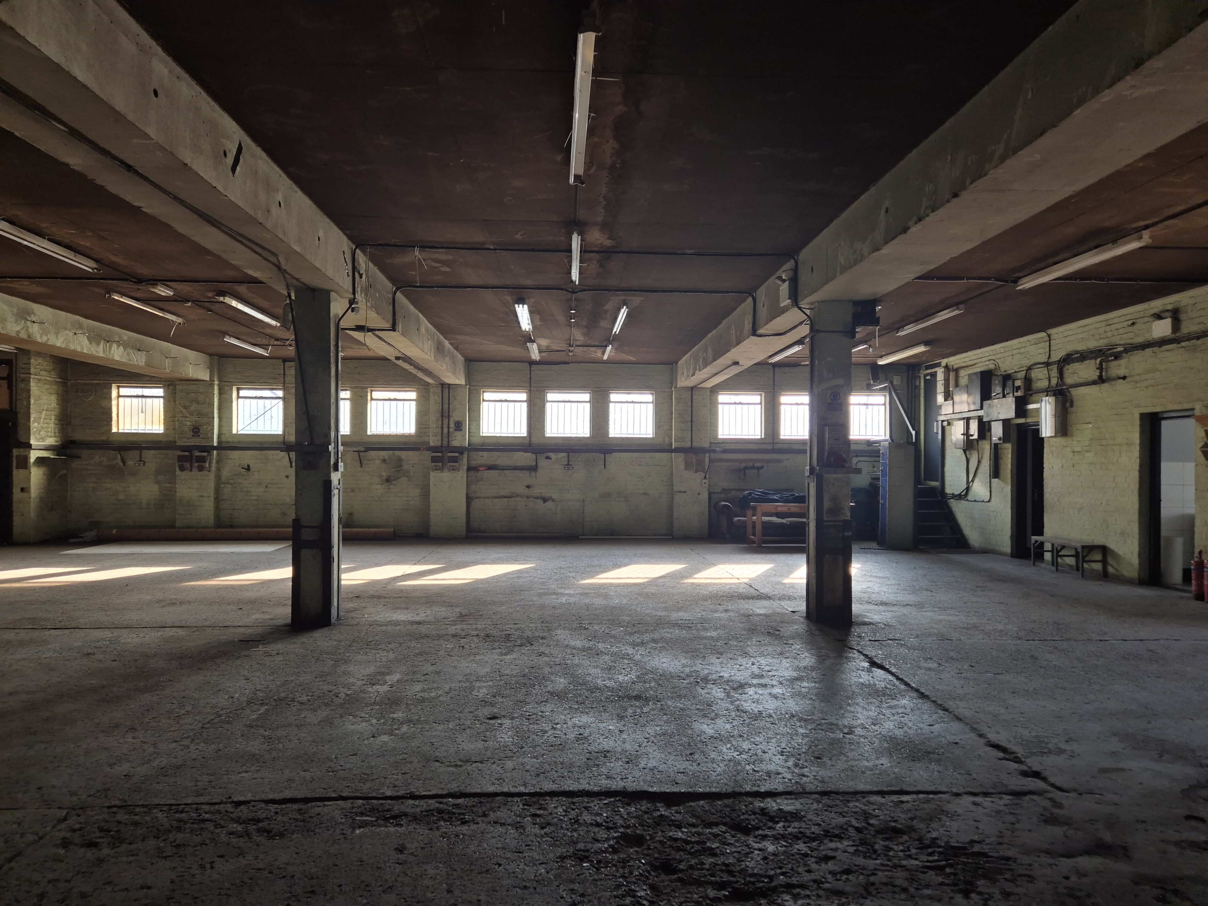 The image shows an empty industrial warehouse with concrete floors and large windows allowing sunlight to stream in.