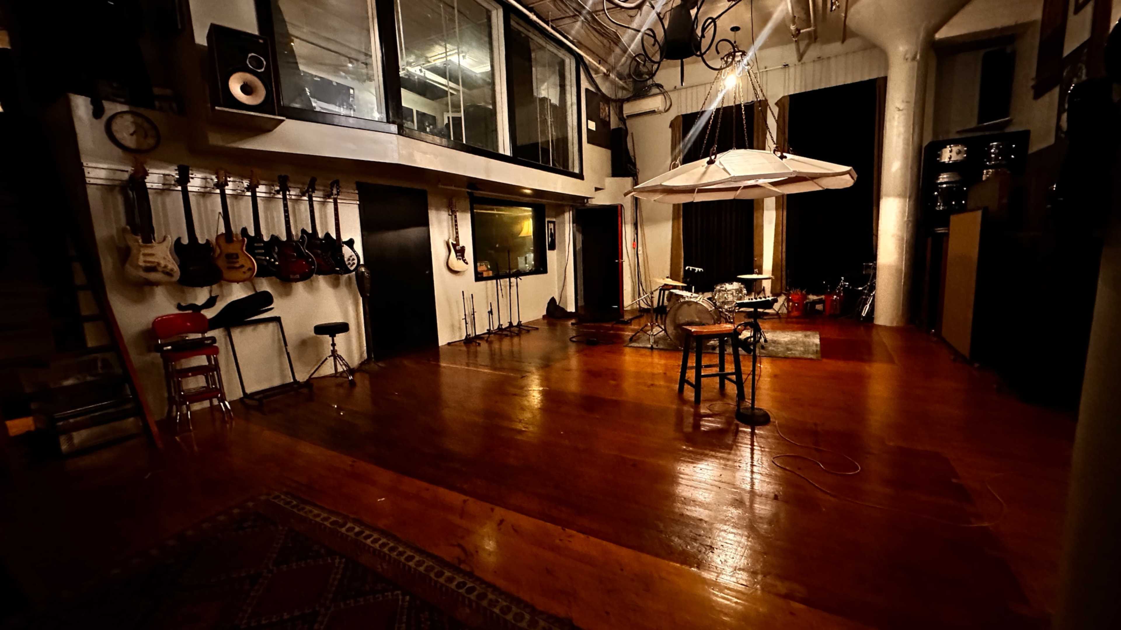 The image shows a spacious music studio with wooden floors, various guitars hanging on the wall, a drum set in the corner, and a central microphone on a stand beneath a hanging light fixture.