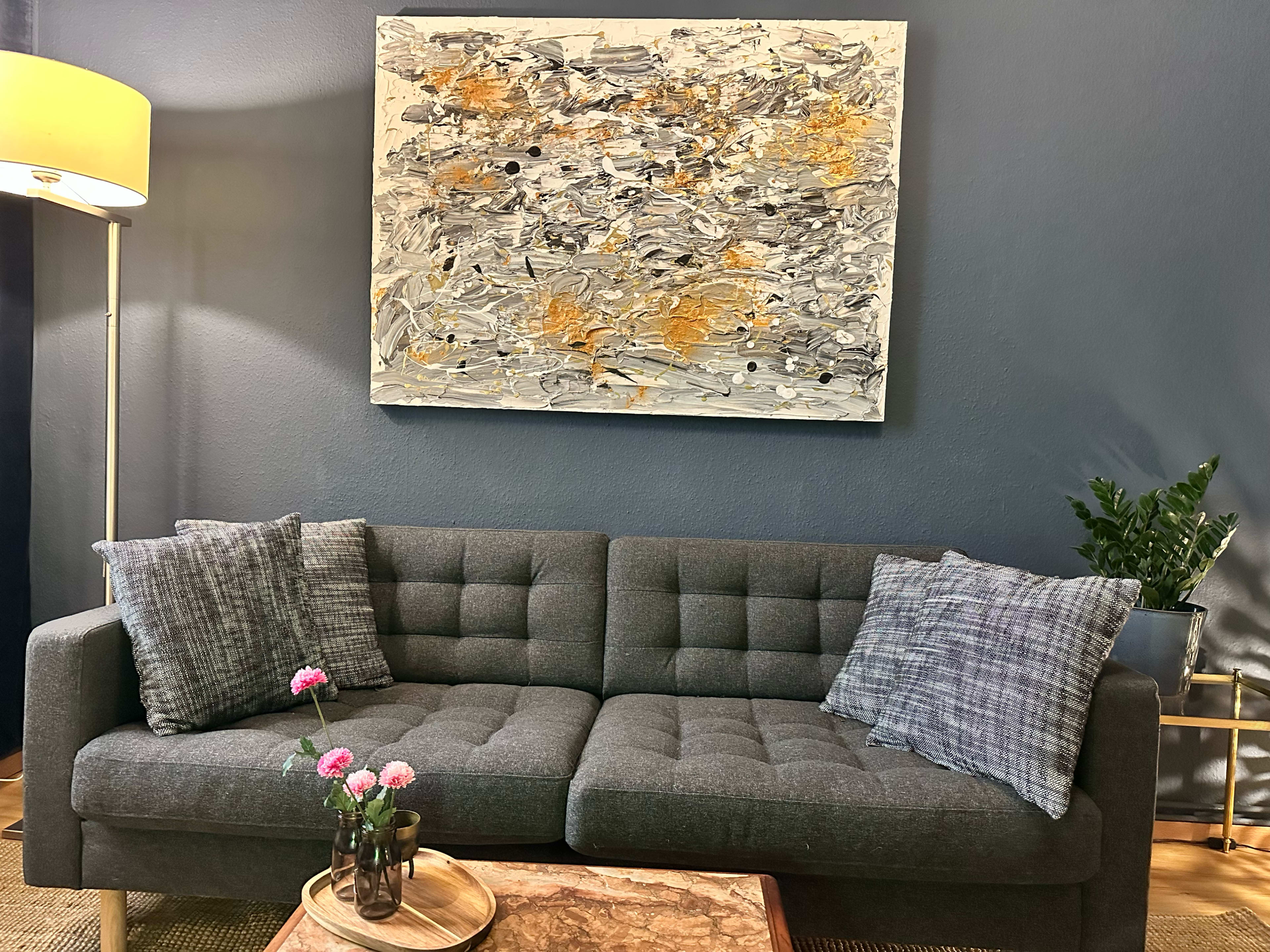 A gray couch with two decorative pillows sits in a living room next to a lamp and a potted plant, beneath an abstract painting on the wall.