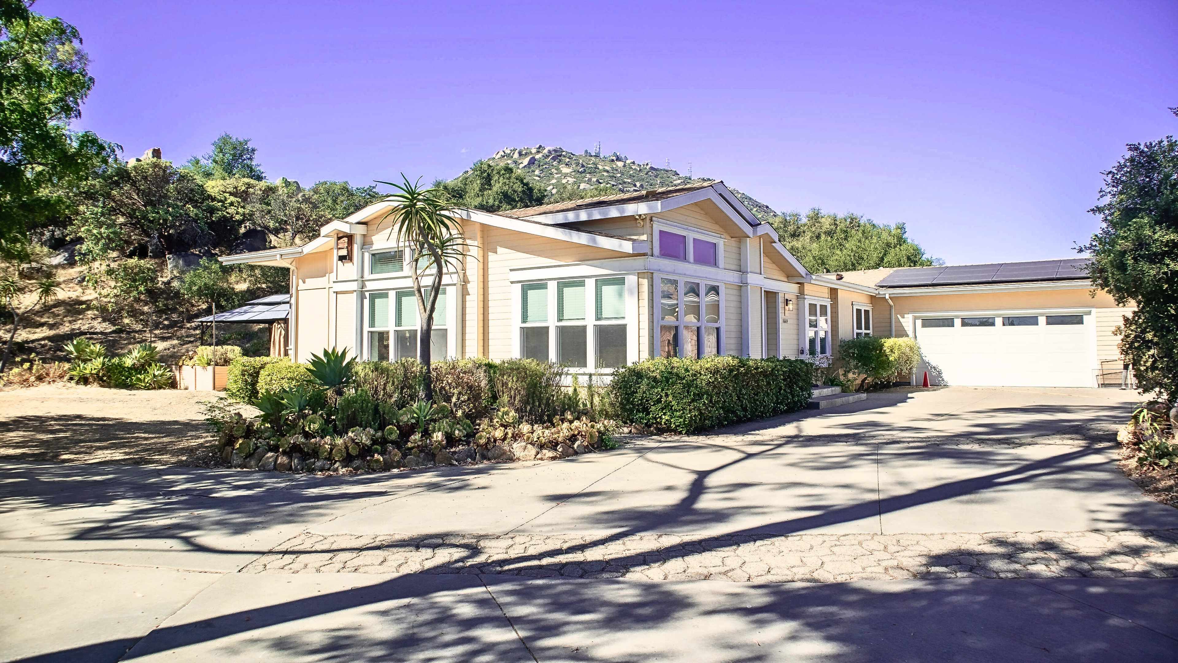 The image shows a single-story house with large windows, surrounded by greenery and a driveway leading to a garage on a hillside.