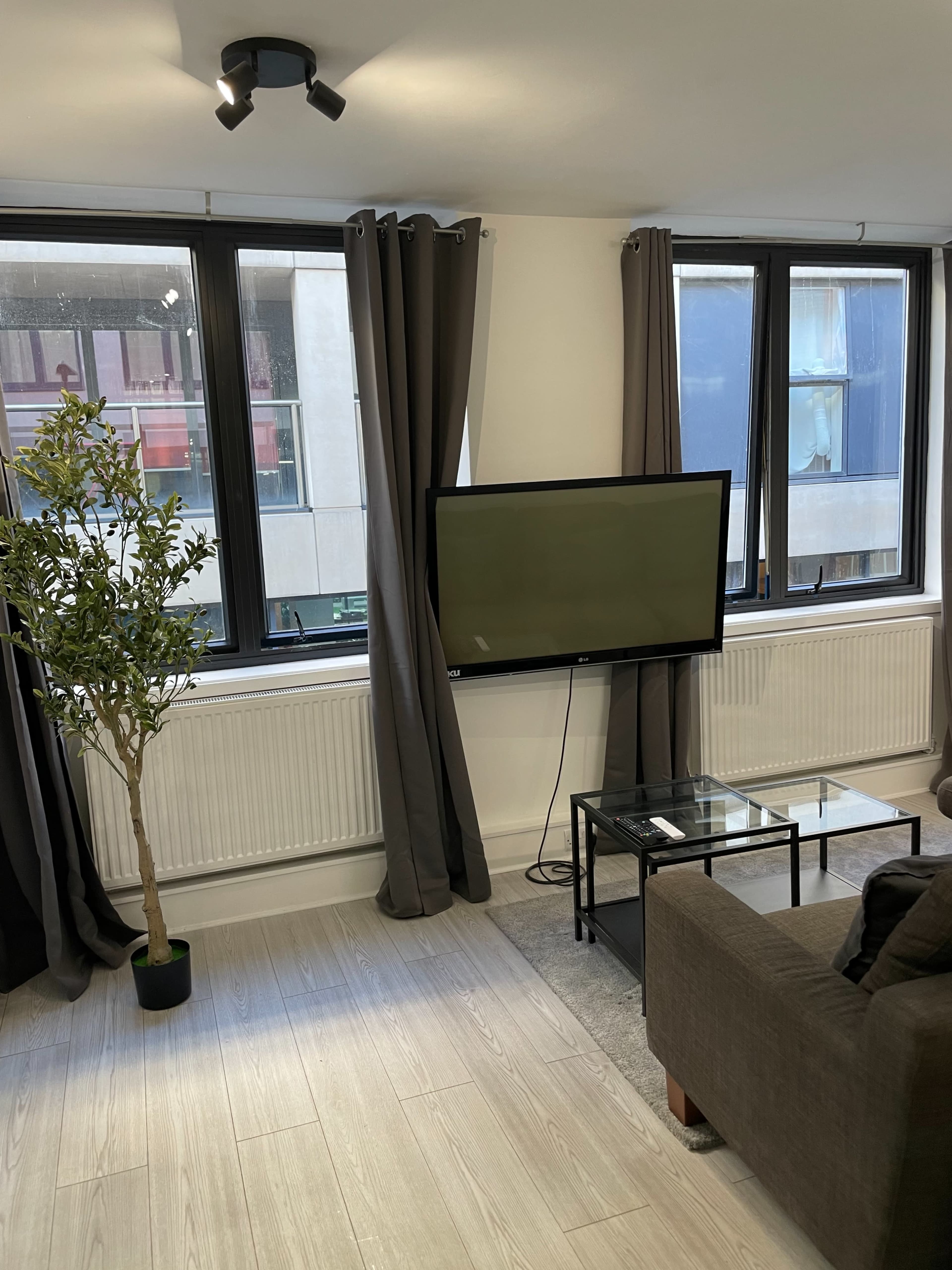 A modern living room features a television mounted on the wall, a glass coffee table, a decorative plant, and large windows dressed with dark curtains.