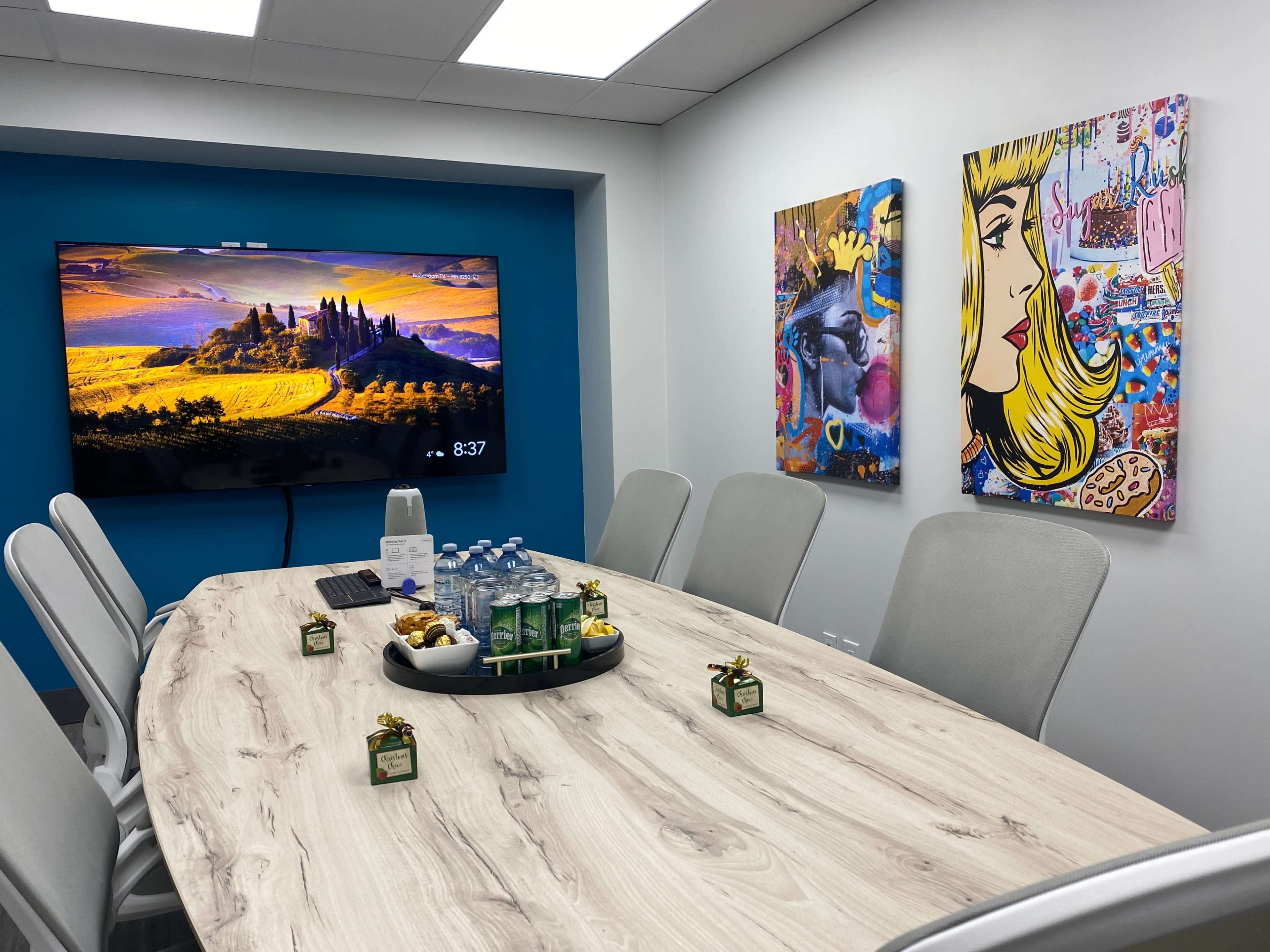 A conference room with a large television, a wooden table set with snacks and beverages, and colorful art on the walls.