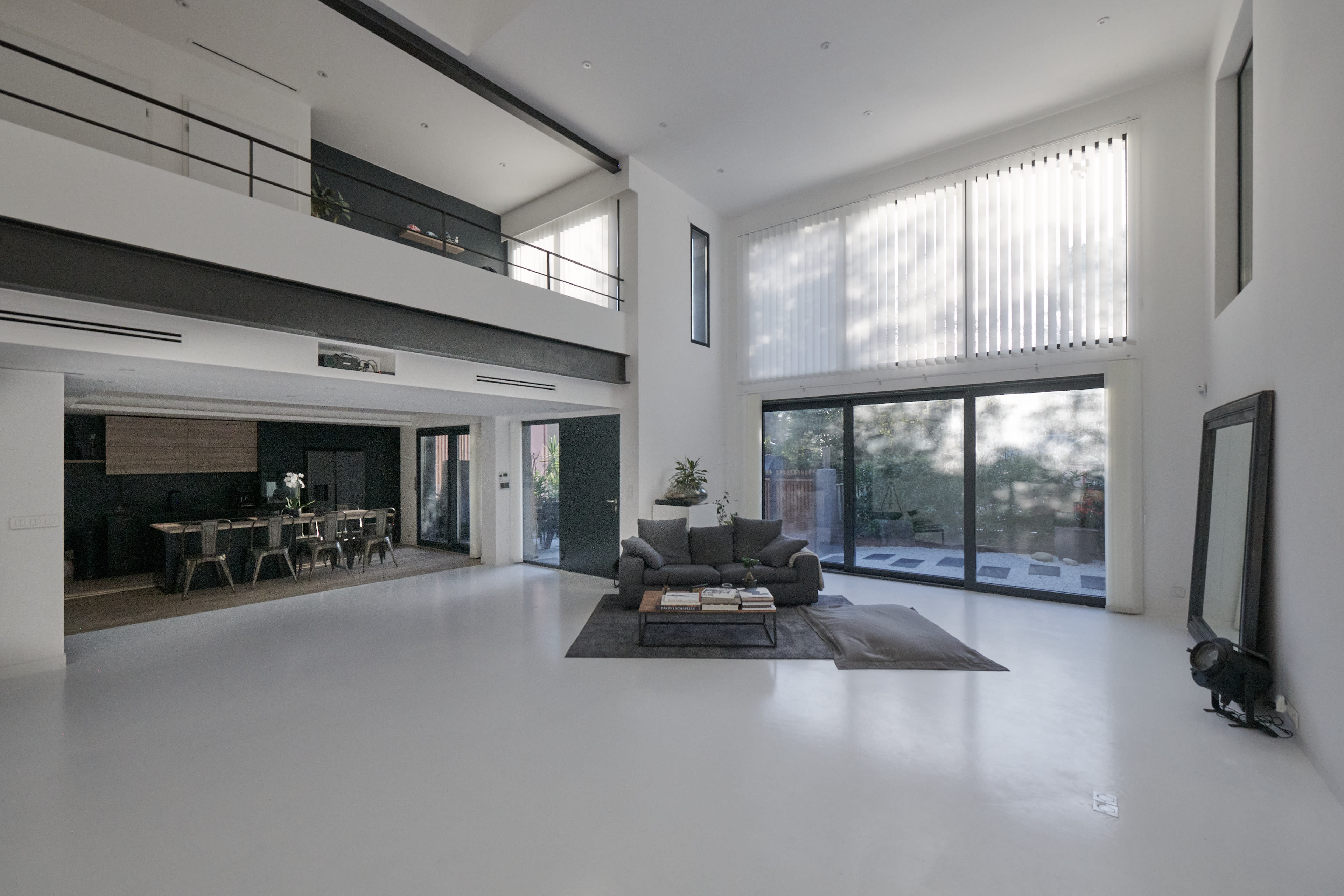 A spacious, modern living area features high ceilings, large windows, minimalist furniture, and an open layout connecting to a dining space.