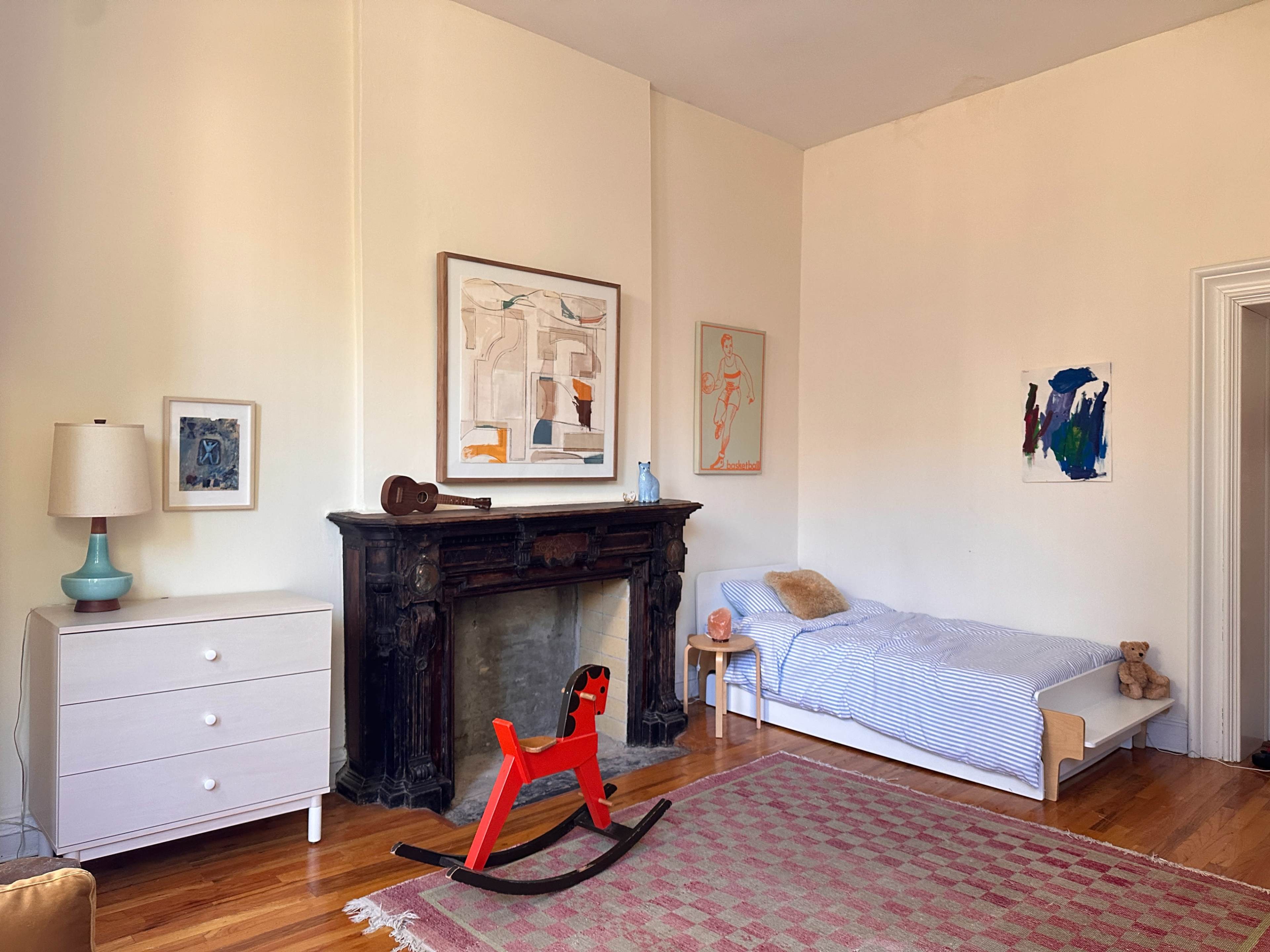 The image features a serene bedroom with a single bed, a wooden rocking horse in the foreground, a dresser with a lamp, and artwork displayed on the walls.
