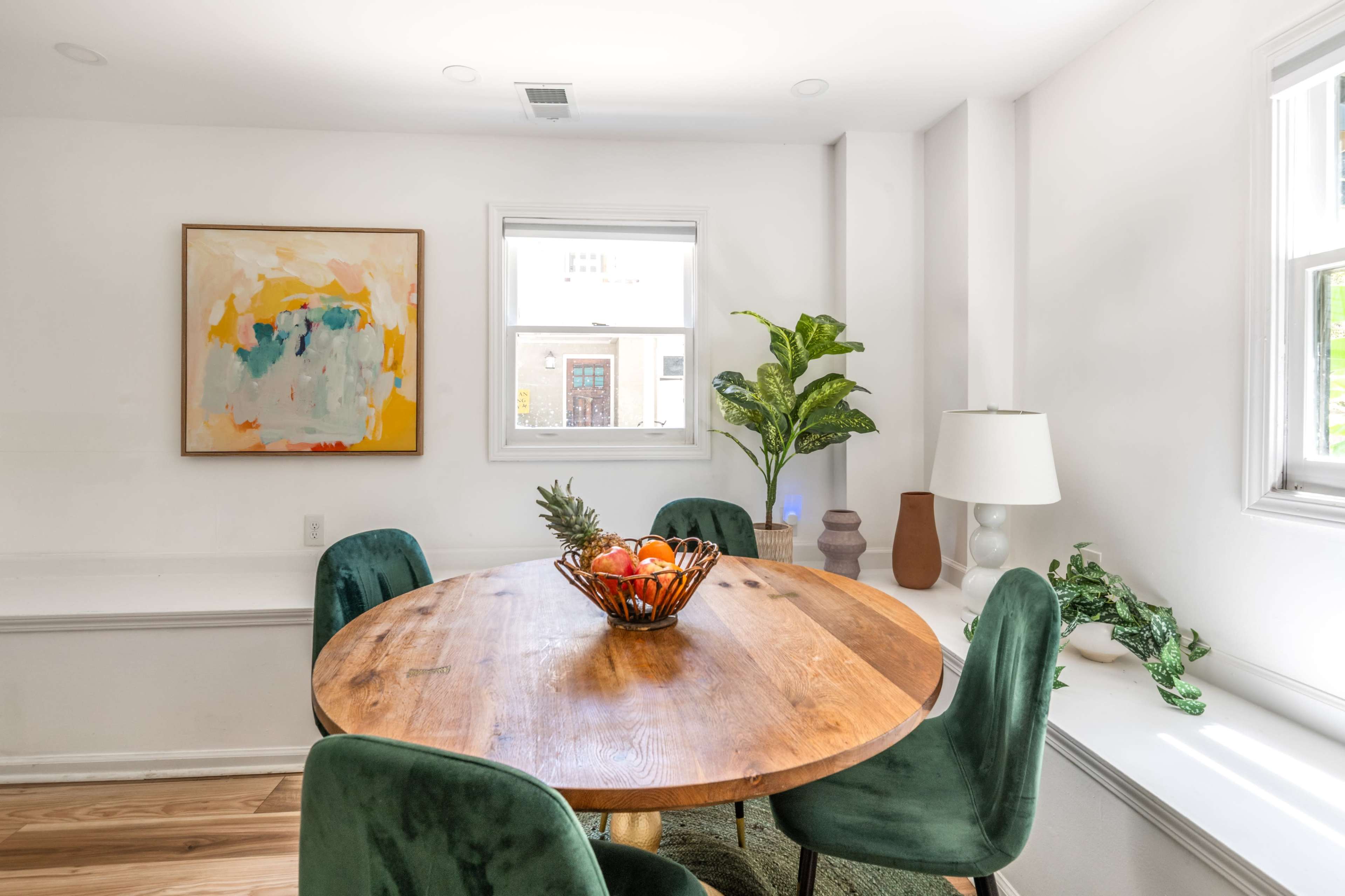 A round wooden table is set in a bright room, surrounded by green velvet chairs, with a fruit bowl at the center and a large abstract painting on the wall.