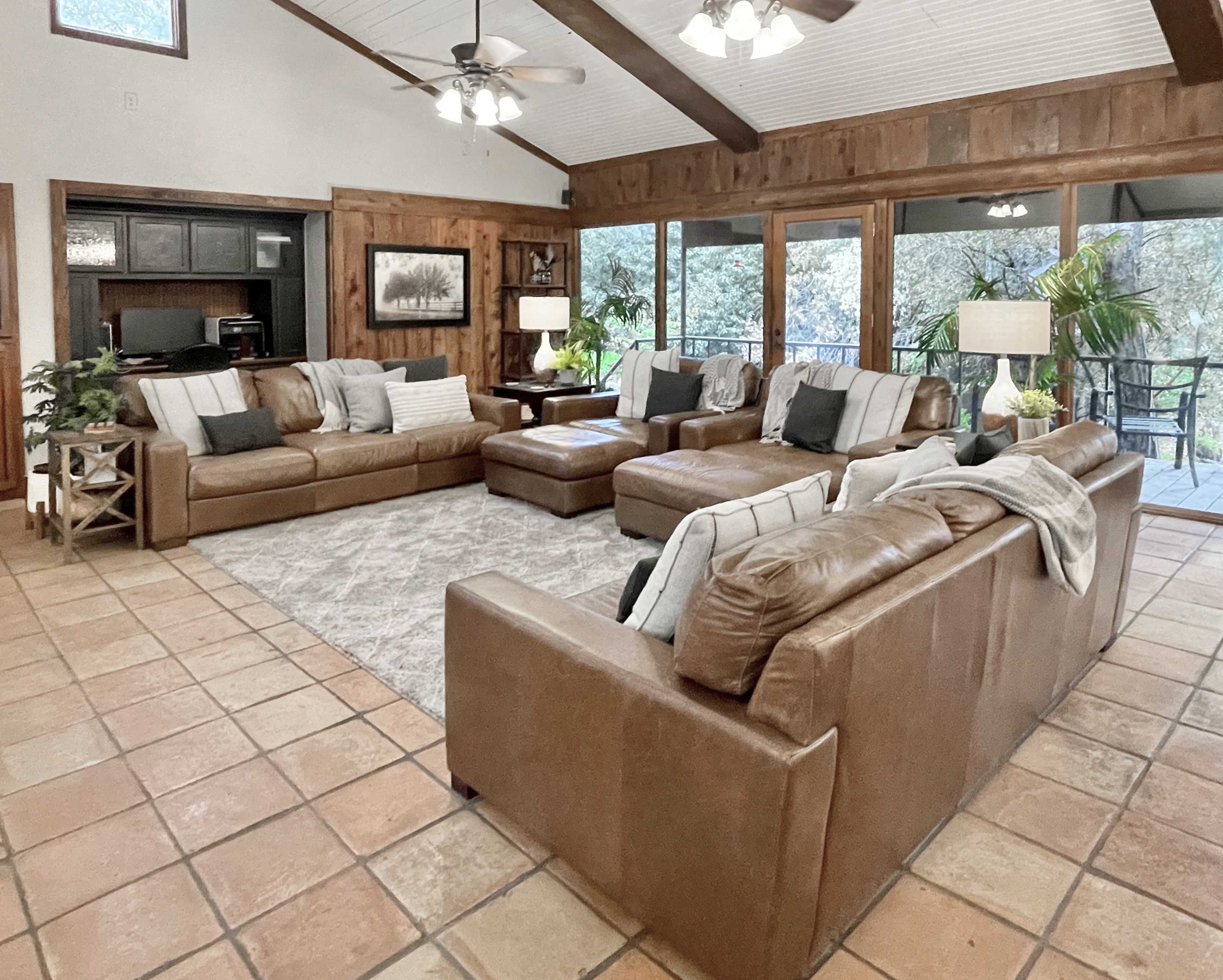 A spacious living room with a large leather sectional sofa, a coffee table, and windows overlooking a green outdoor area.