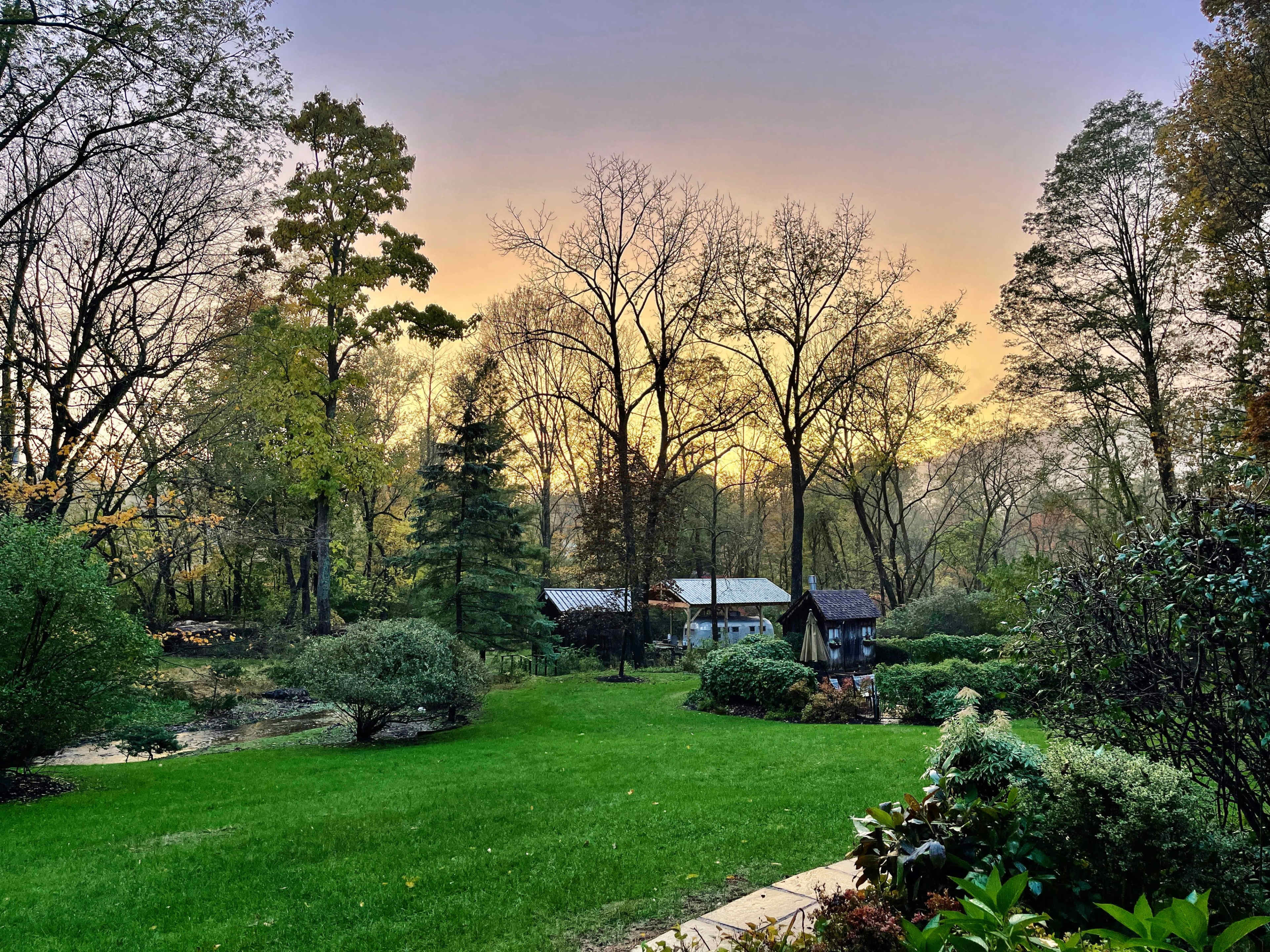 A well-maintained garden with green grass, bushes, and trees, set against a background of wooden structures and a colorful sunset sky.