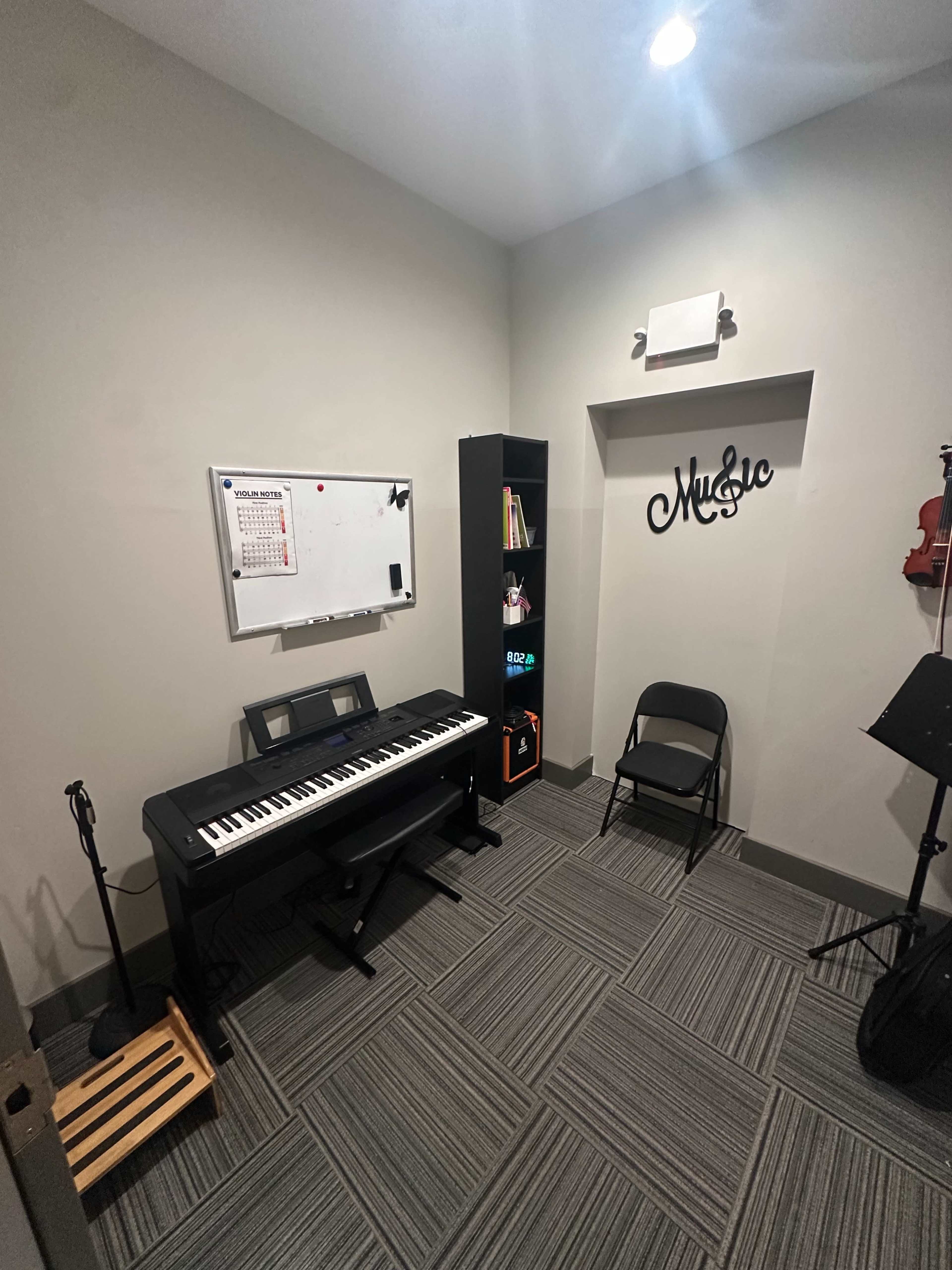 A small music practice room features a digital piano, a bookshelf with various items, a calendar on the wall, and a charred black chair.