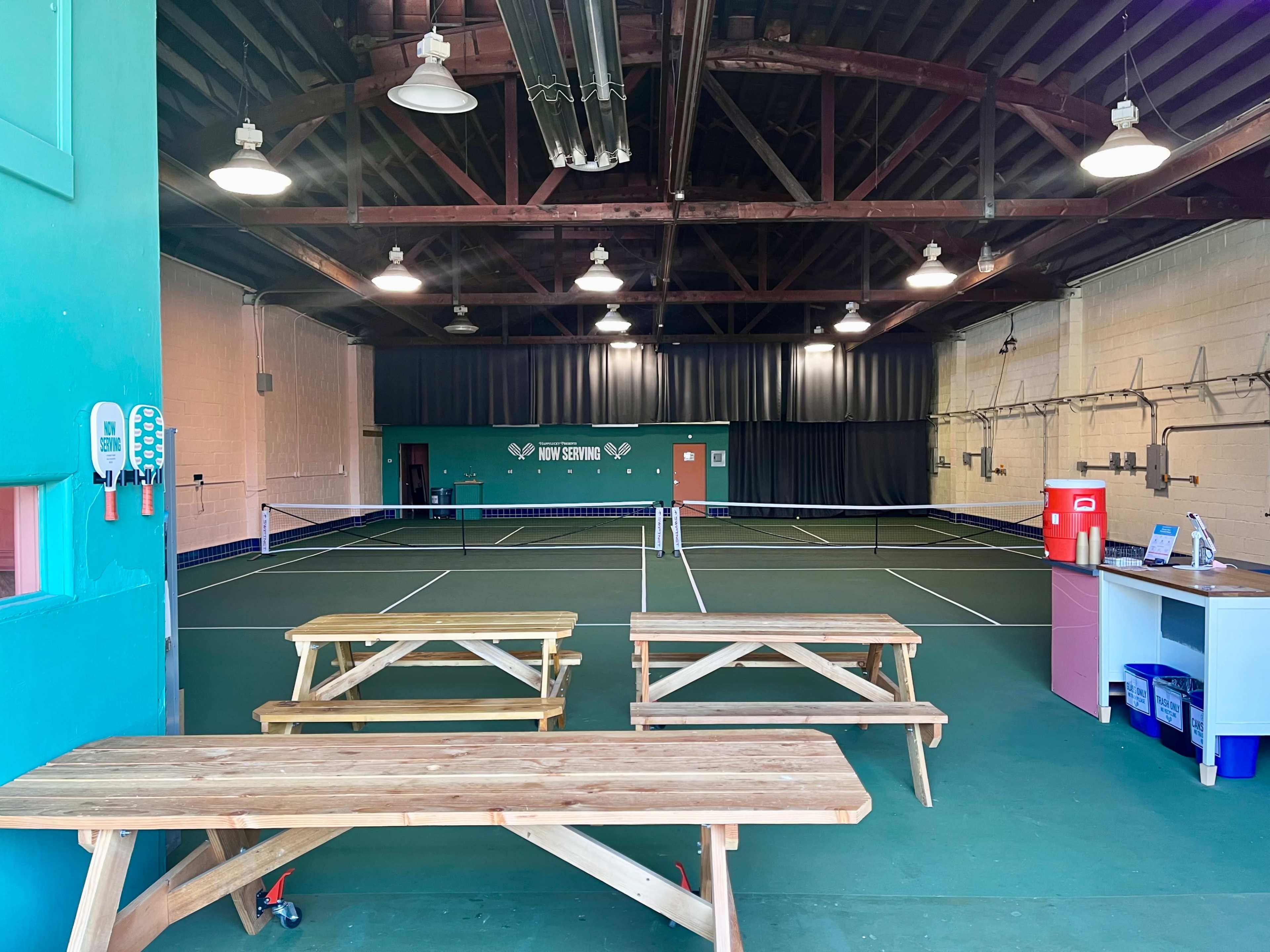 The image shows an indoor tennis facility featuring two courts, wooden benches, and a serving area with a water cooler and a small table.
