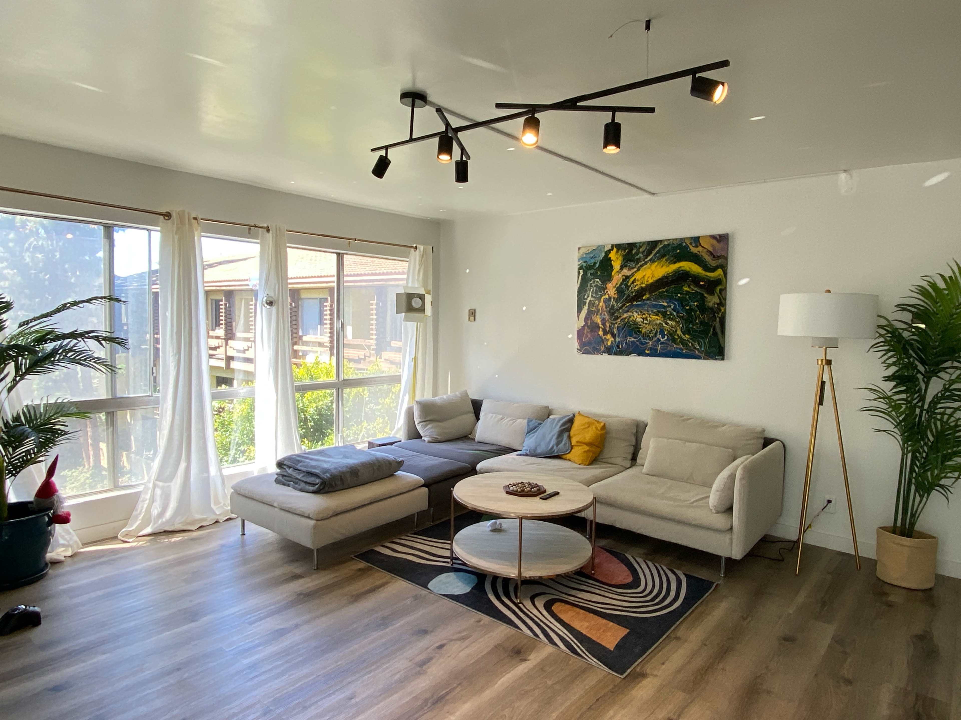The living room features a light-colored sectional sofa, a round coffee table, and large windows that let in natural light, with plants and decorative artwork enhancing the space.