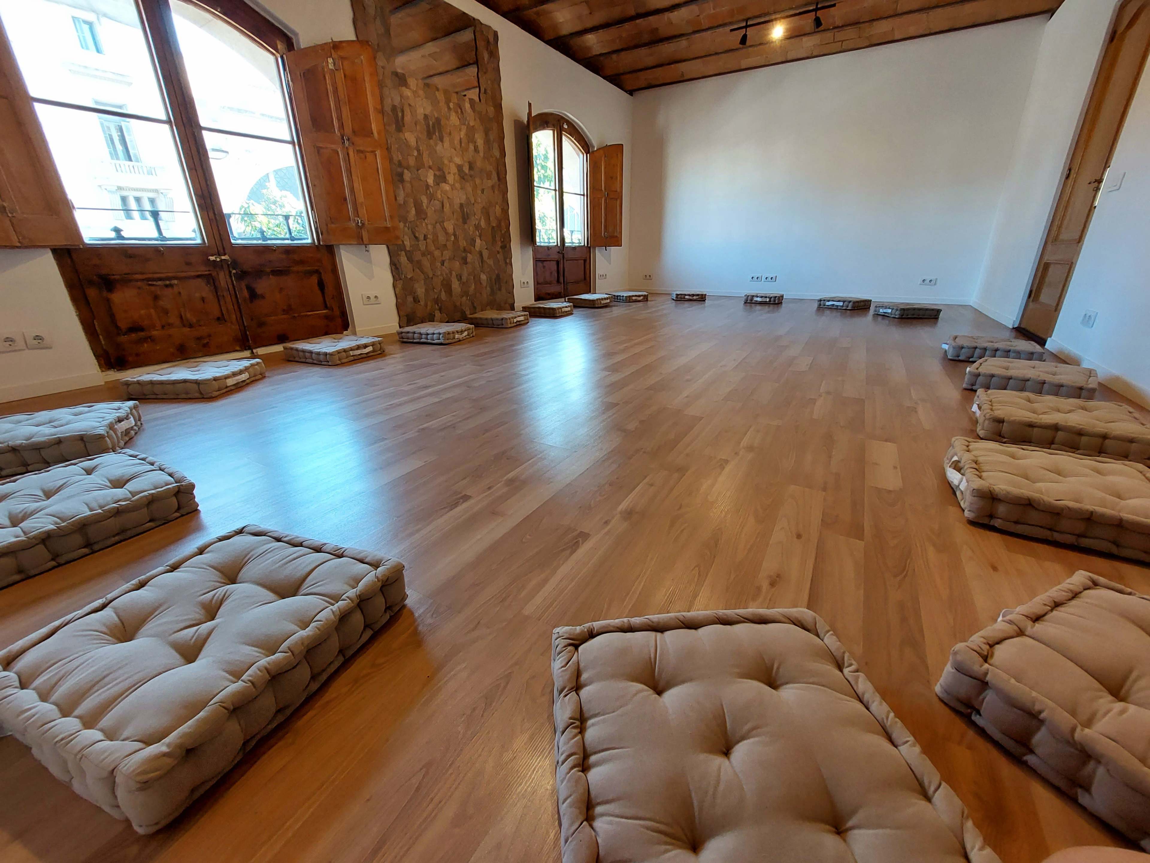 The image shows a wooden-floored room with several cushioned mats arranged on the floor and large windows with shutters.