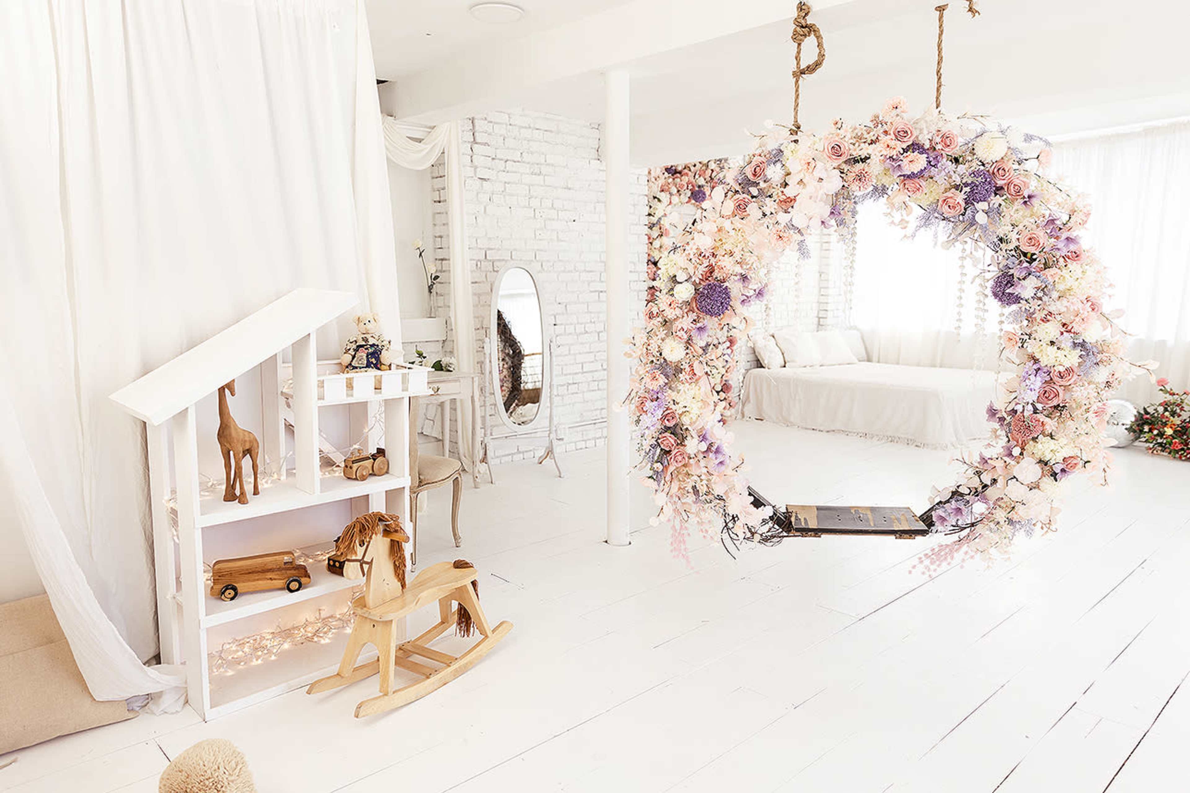 A bright, airy room with a floral swing, a white wooden playhouse, and a rocking horse.