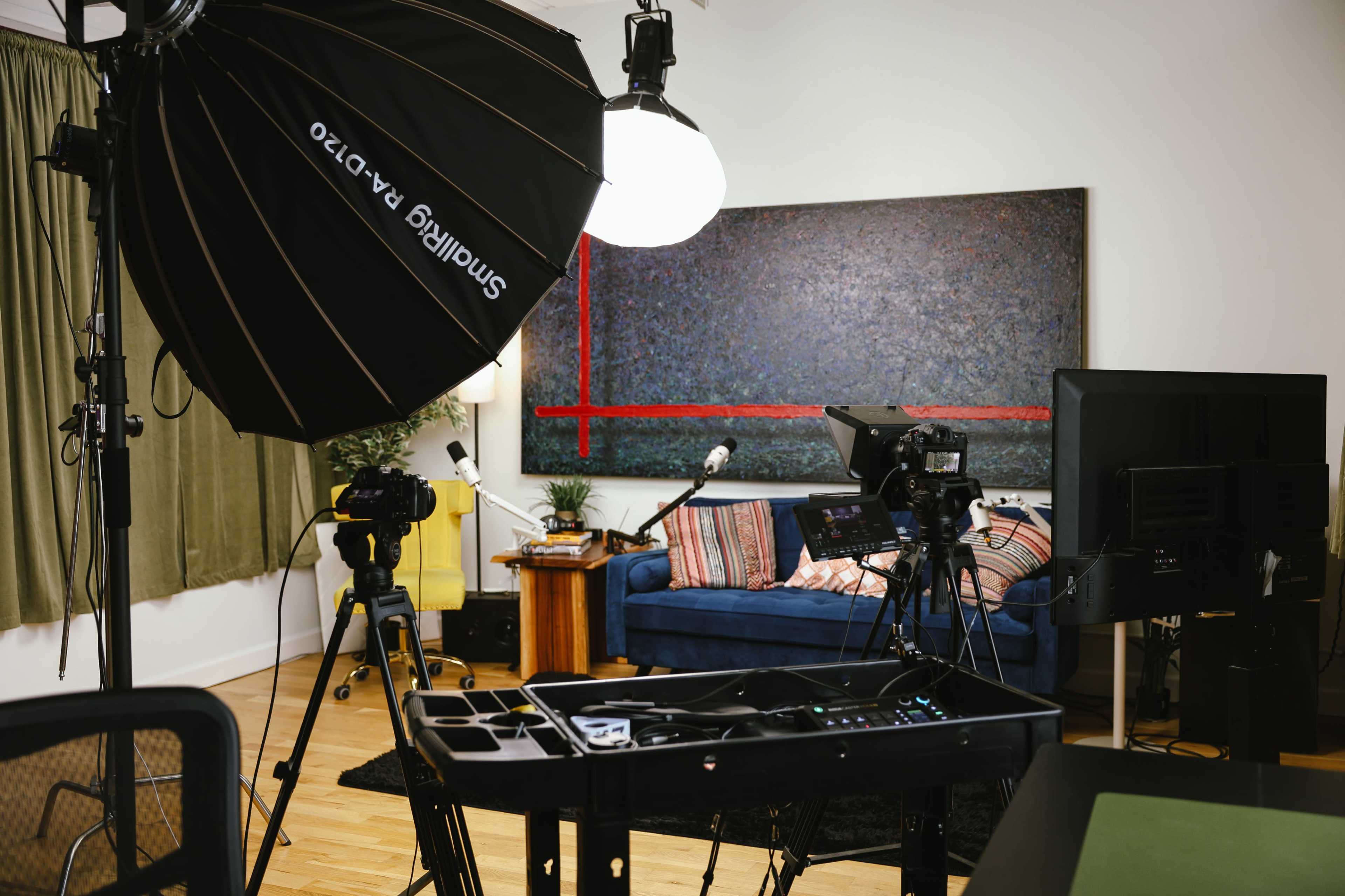The image shows a media production setup in a studio with camera equipment, lighting, a couch, and a table with microphones.