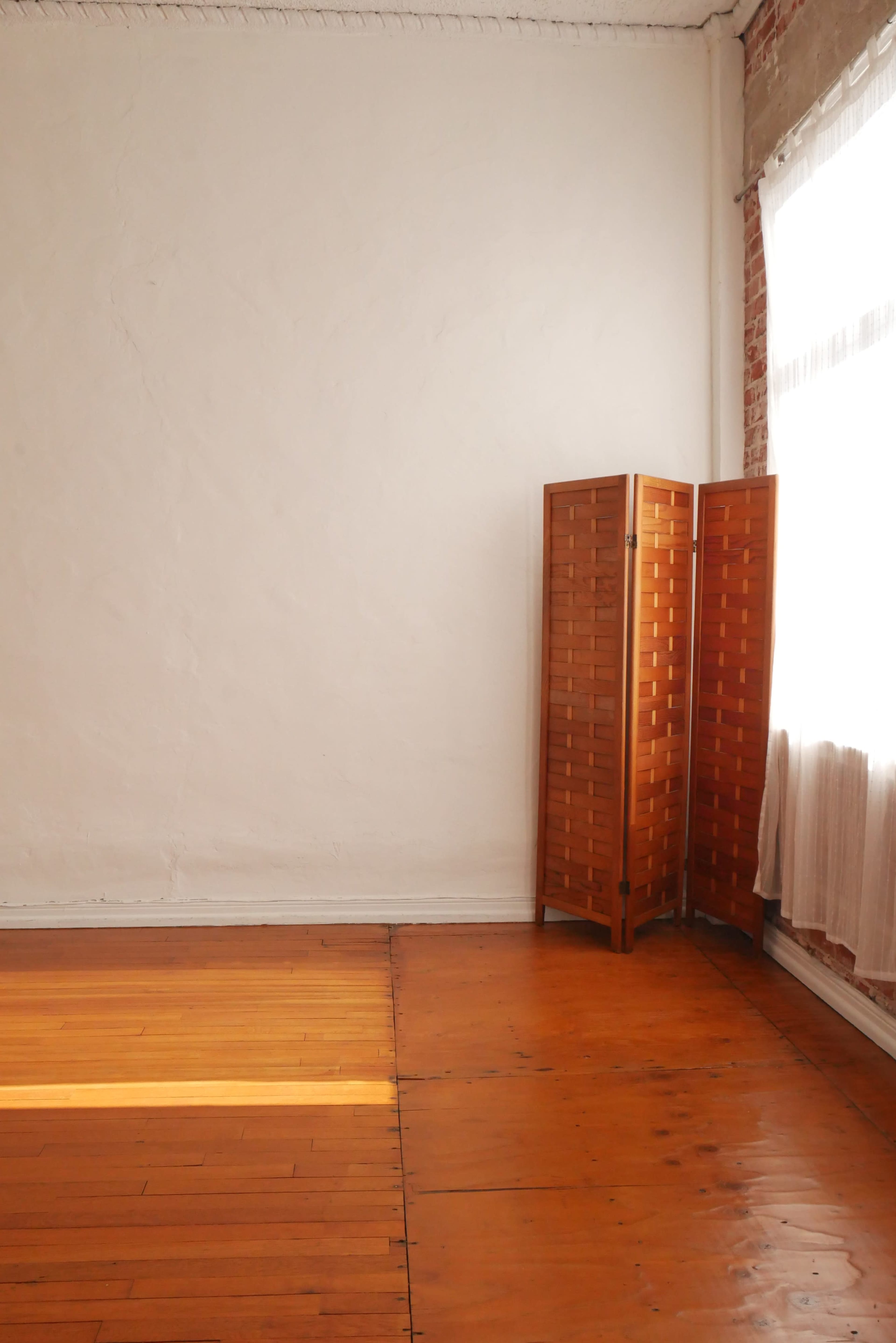 A spacious room with bare walls, polished wooden floors, and a wooden folding screen in the corner near a window with sheer curtains.