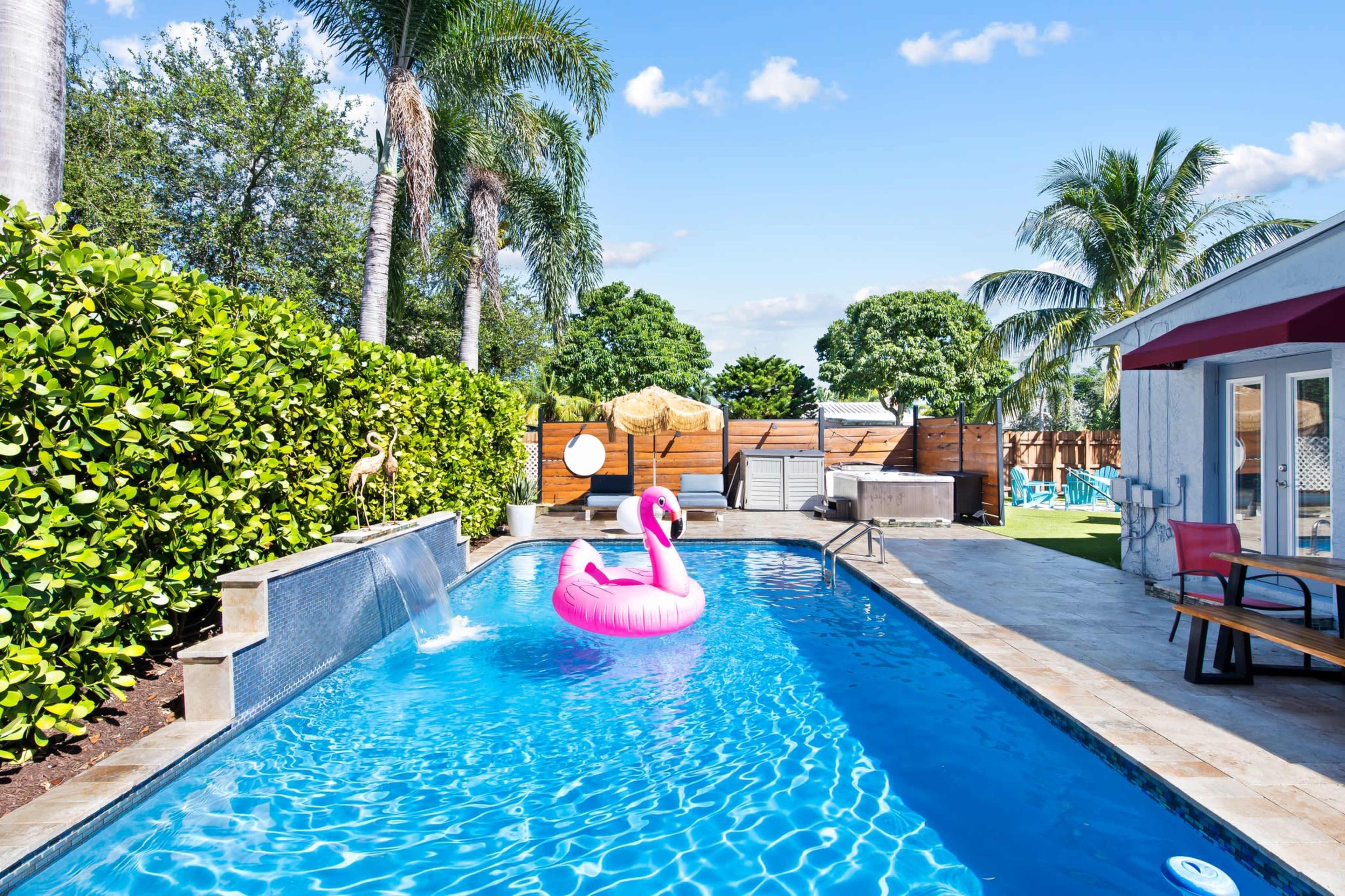 A swimming pool with a pink flamingo float, surrounded by palm trees and green hedges under a clear blue sky.