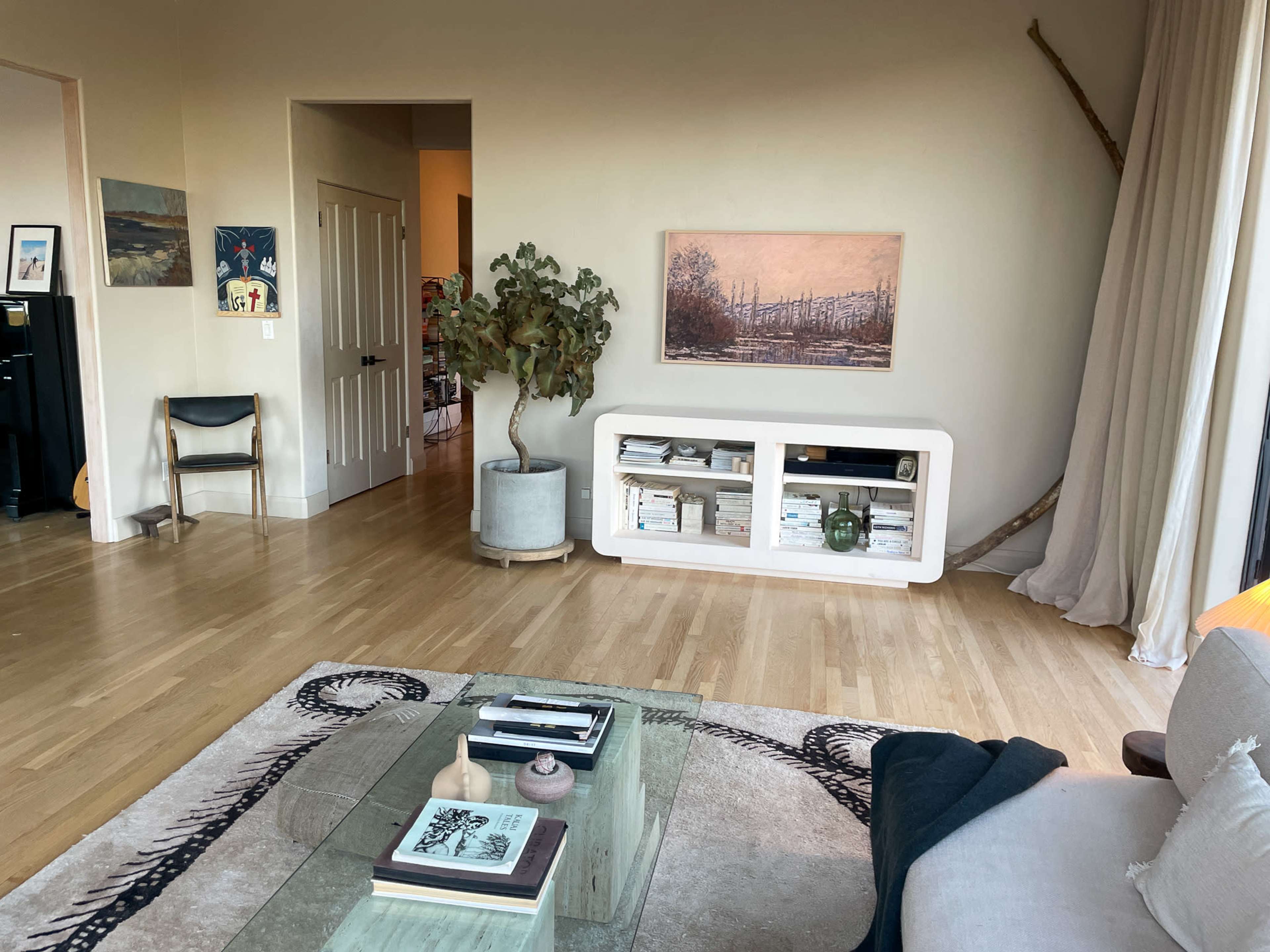 The image depicts a modern living room with wooden flooring, a large window, a plant beside a low white shelf filled with books, and a neutral-toned artwork hanging on the wall.