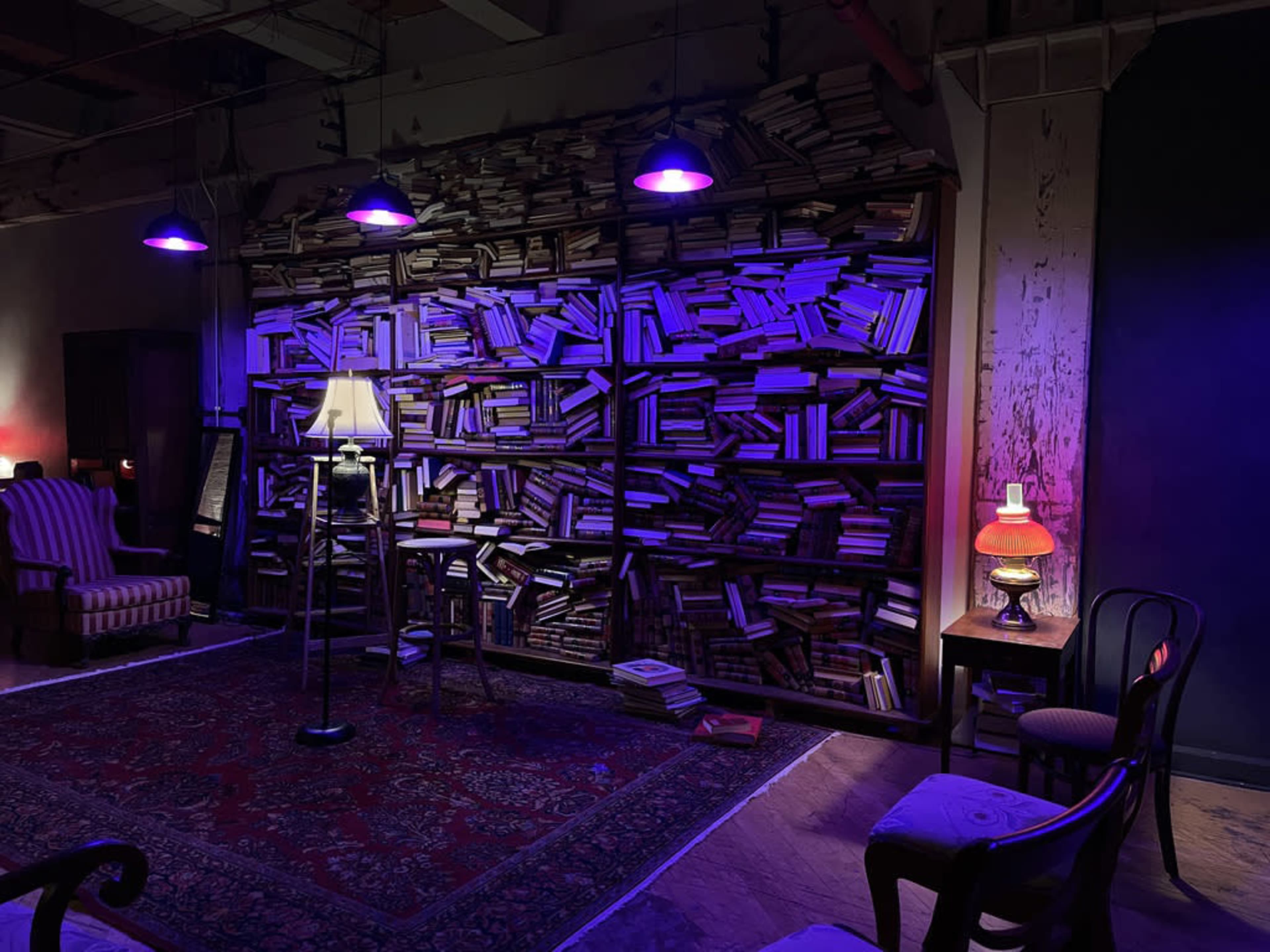 A dimly lit room features a large bookshelf filled with books, a table with a lamp, and various chairs arranged around a patterned rug.