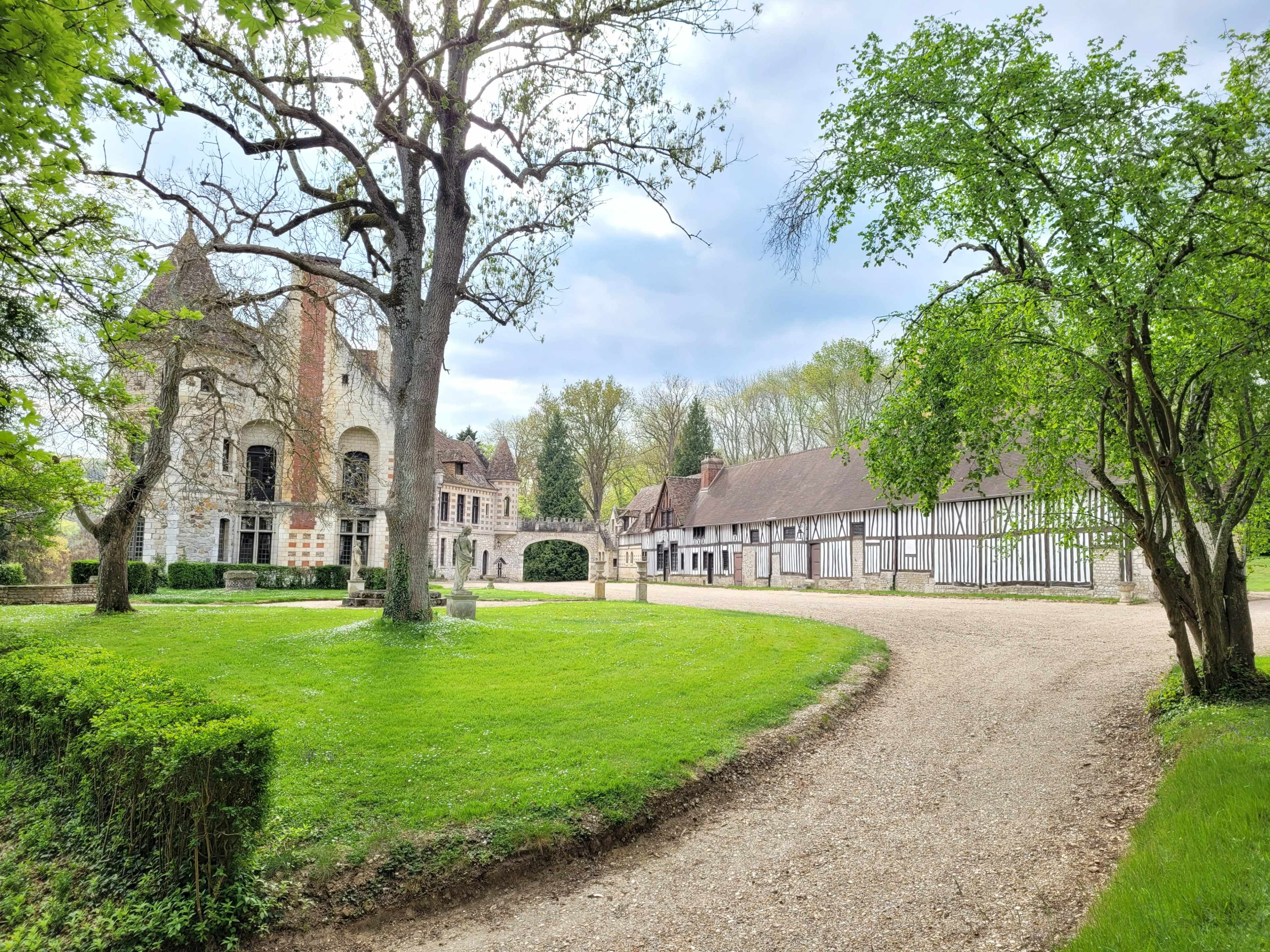 A gravel pathway leading through a lush green landscape, bordered by trees and a historic stone building with a half-timbered structure in the background.