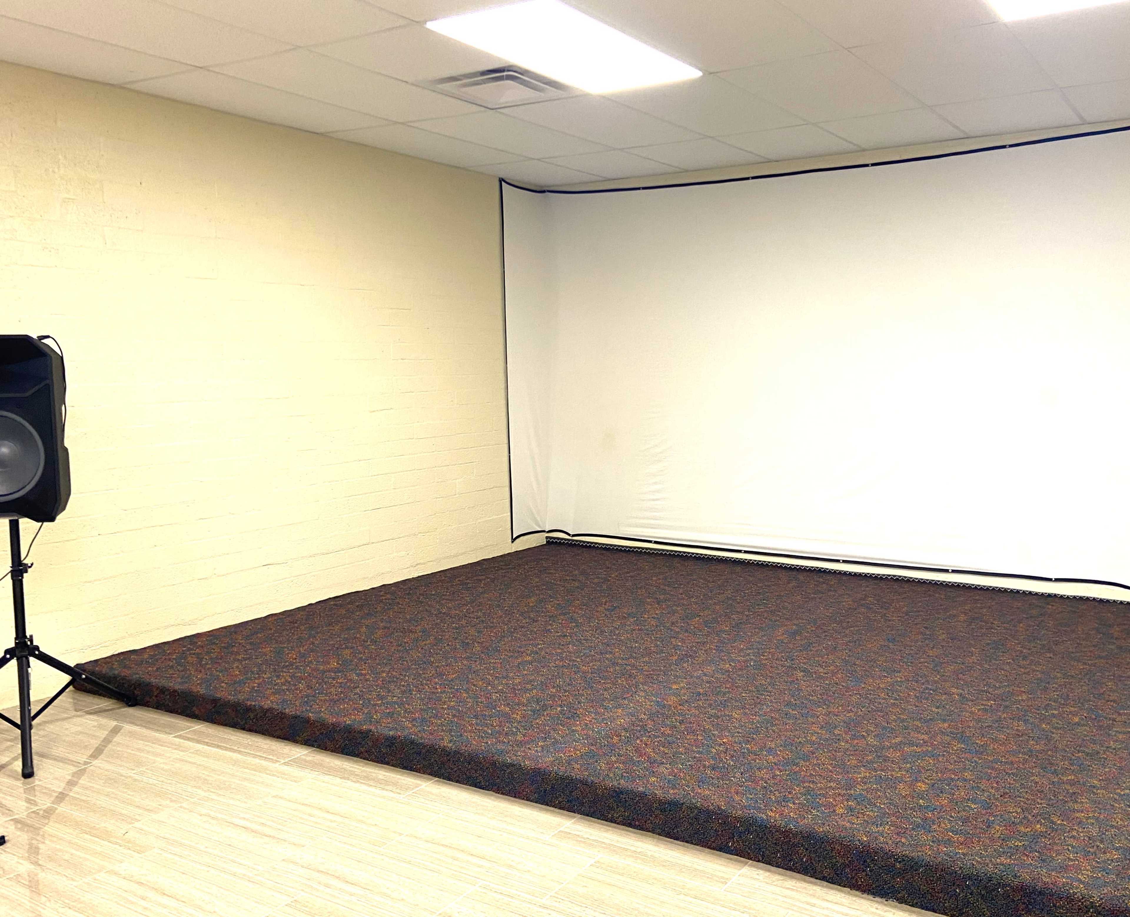 An empty rehearsal space with a large, flat carpeted area and a white backdrop against the wall.