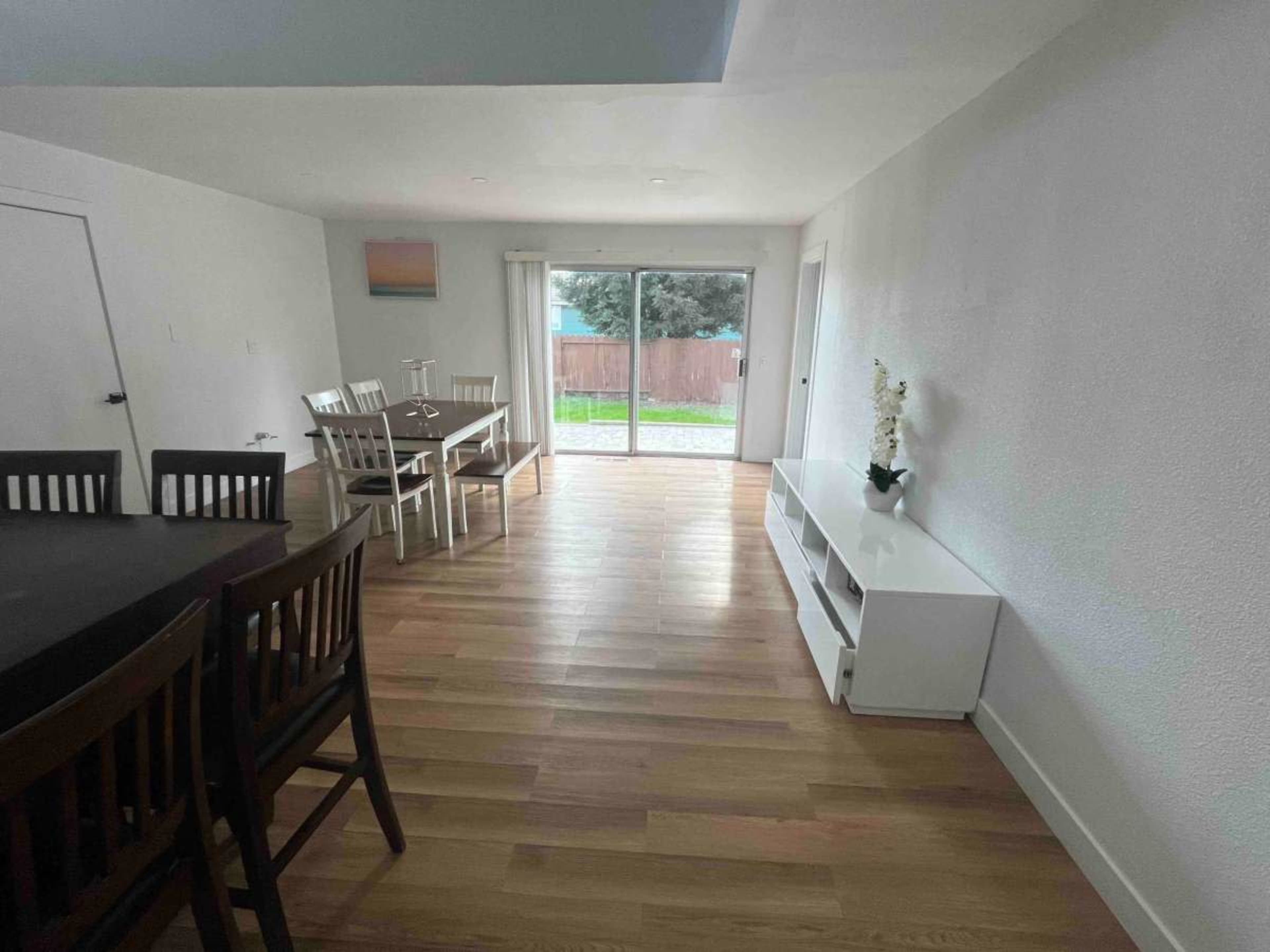 The image shows a modern interior space with a dining area featuring a table and chairs, a white media console, and a sliding glass door leading to an outdoor area.