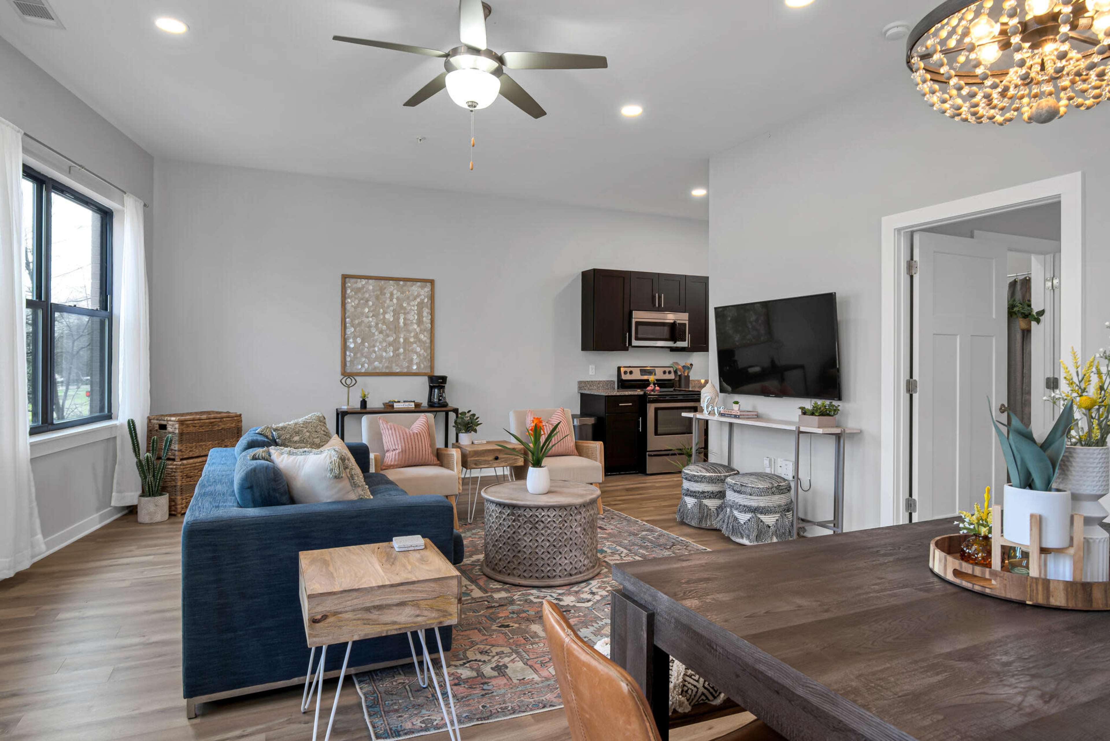 A modern living room with a blue sectional sofa, a small wooden table, and a dining area, featuring a large window and a well-equipped kitchen in the background.