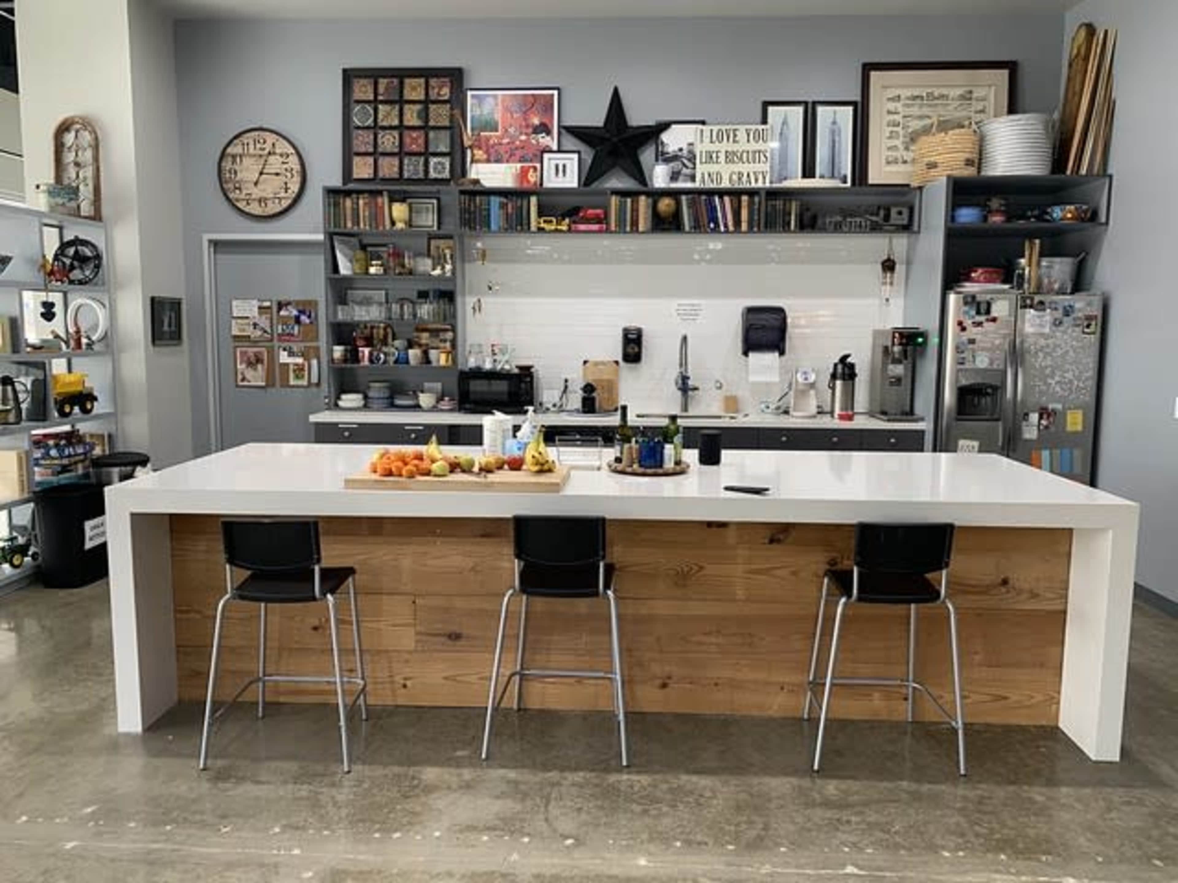 A modern kitchen with a large white island and wooden front, featuring a variety of fruit on top and shelves filled with kitchenware in the background.