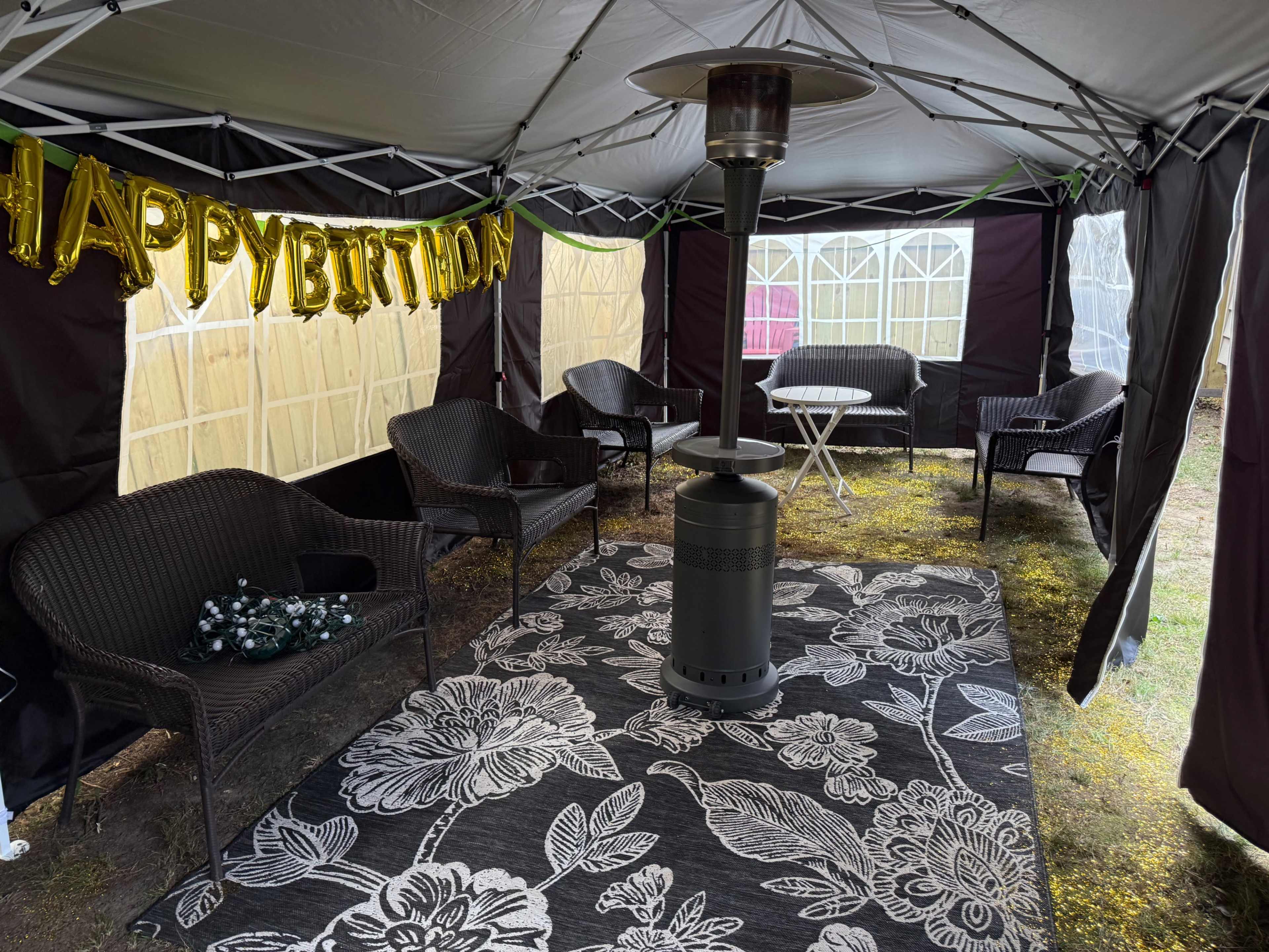 A canopy tent is set up with a "HAPPY BIRTHDAY" banner, surrounded by chairs and a patio heater on a patterned rug.