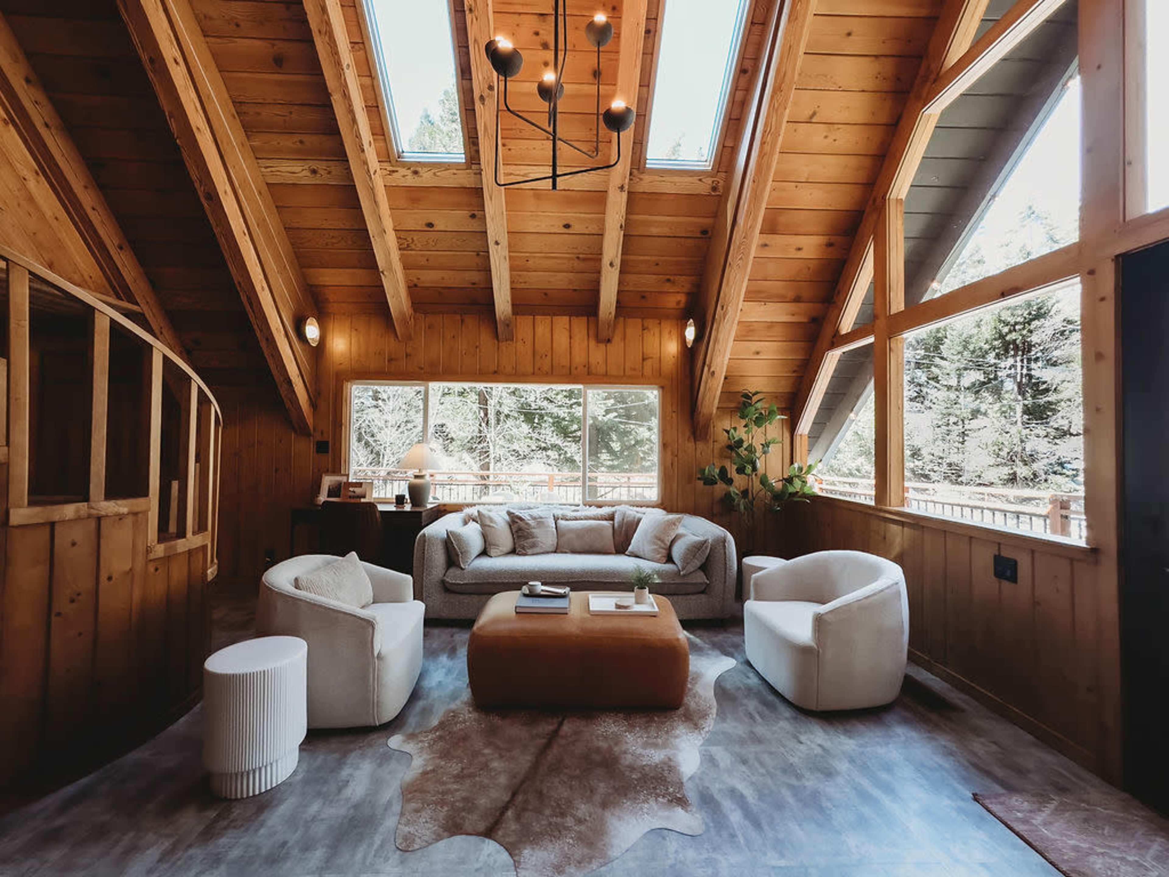 The image shows a cozy living room with wooden walls, a vaulted ceiling, large windows, and modern furnishings.