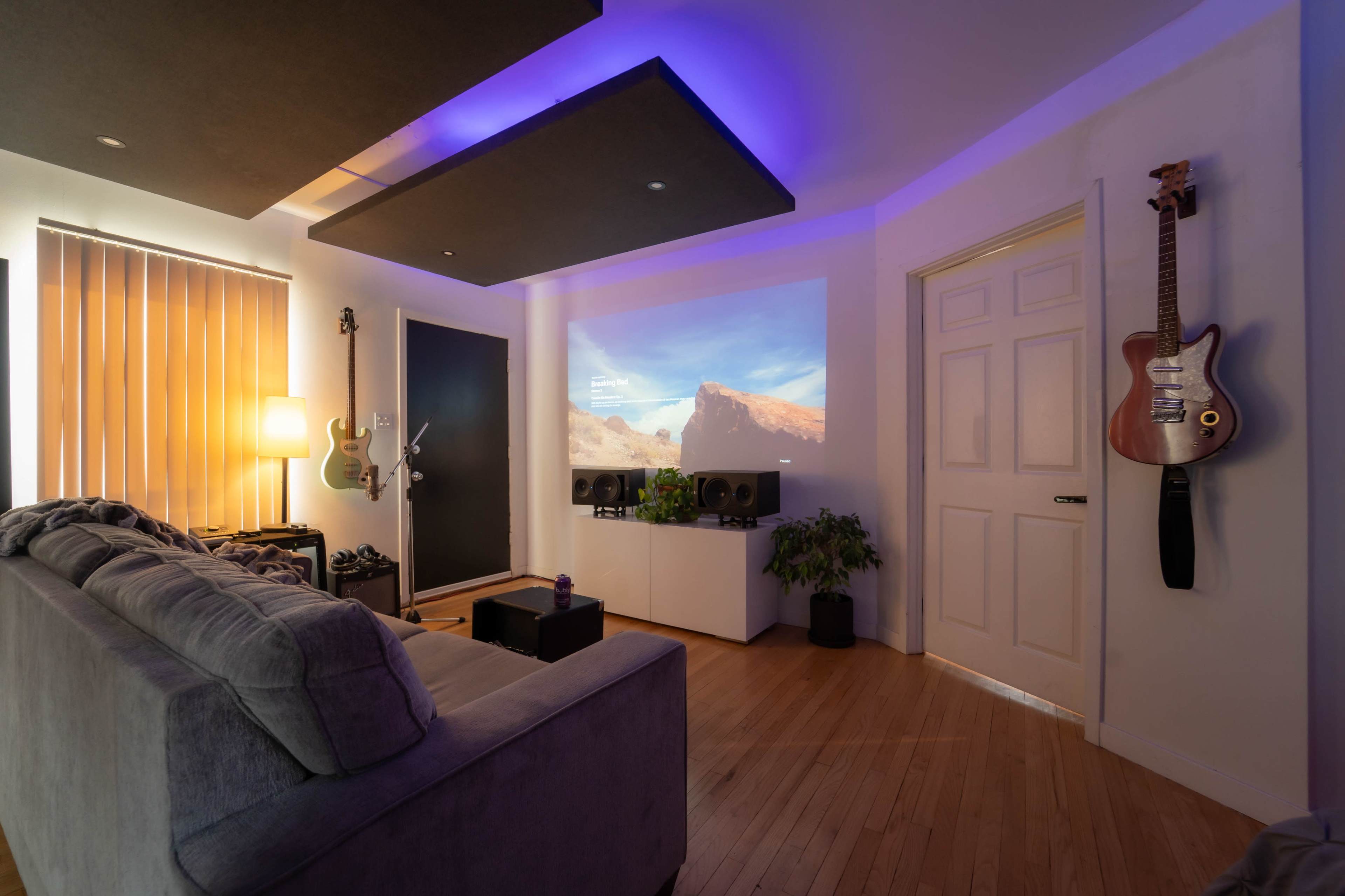 A modern living room with a projector displaying a landscape scene, a grey sofa, and a guitar hanging on the wall.